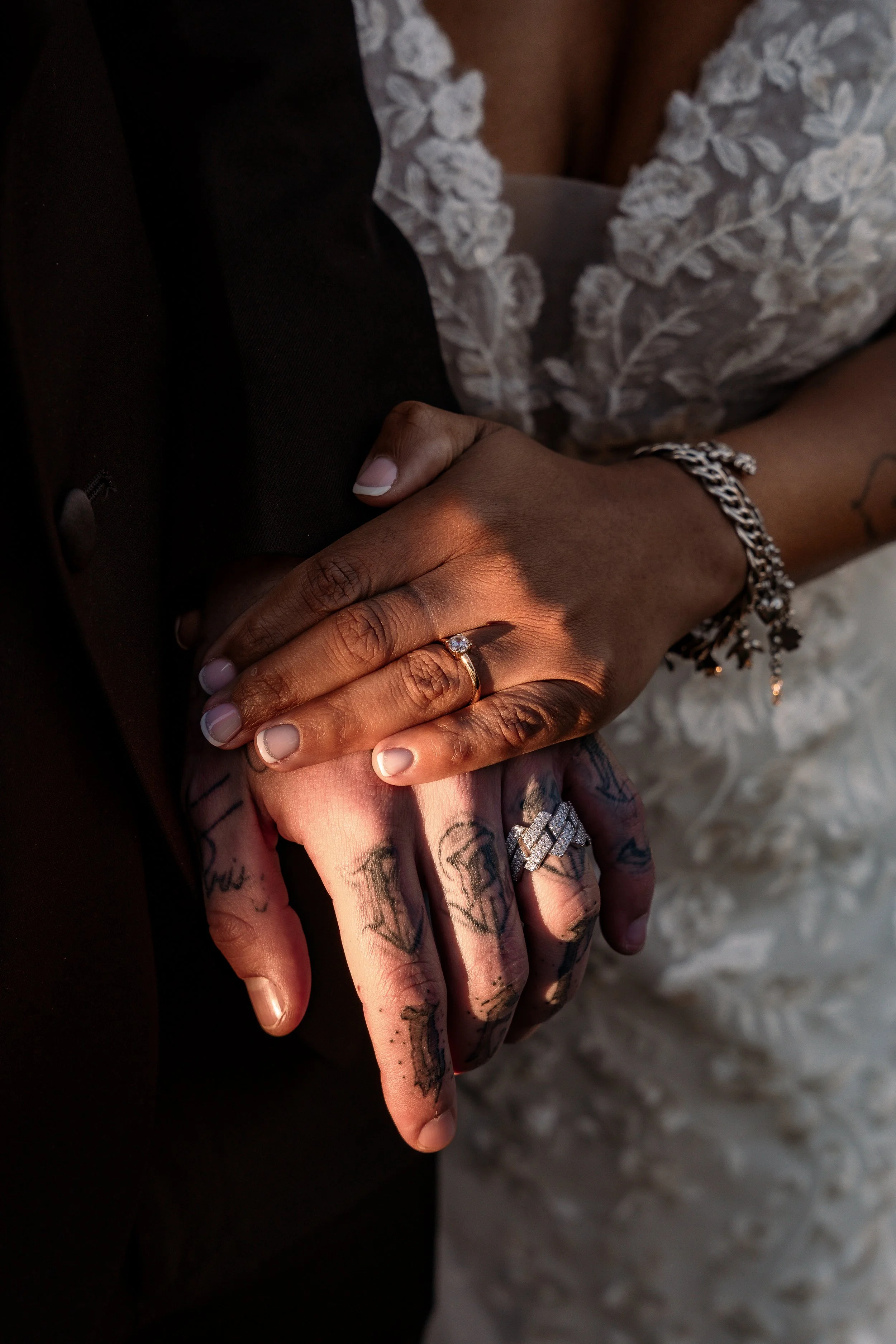 Two hands, one with a diamond engagement ring, clasped together. The person with the rings has tattoos on their fingers and is wearing a silver bracelet. The background shows part of a wedding dress.