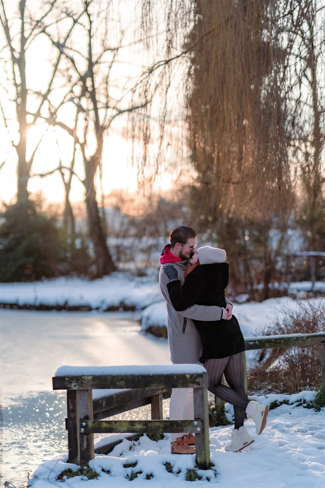 A couple embracing outdoors on a snowy winter day at sunset, near a frozen pond and leafless trees.