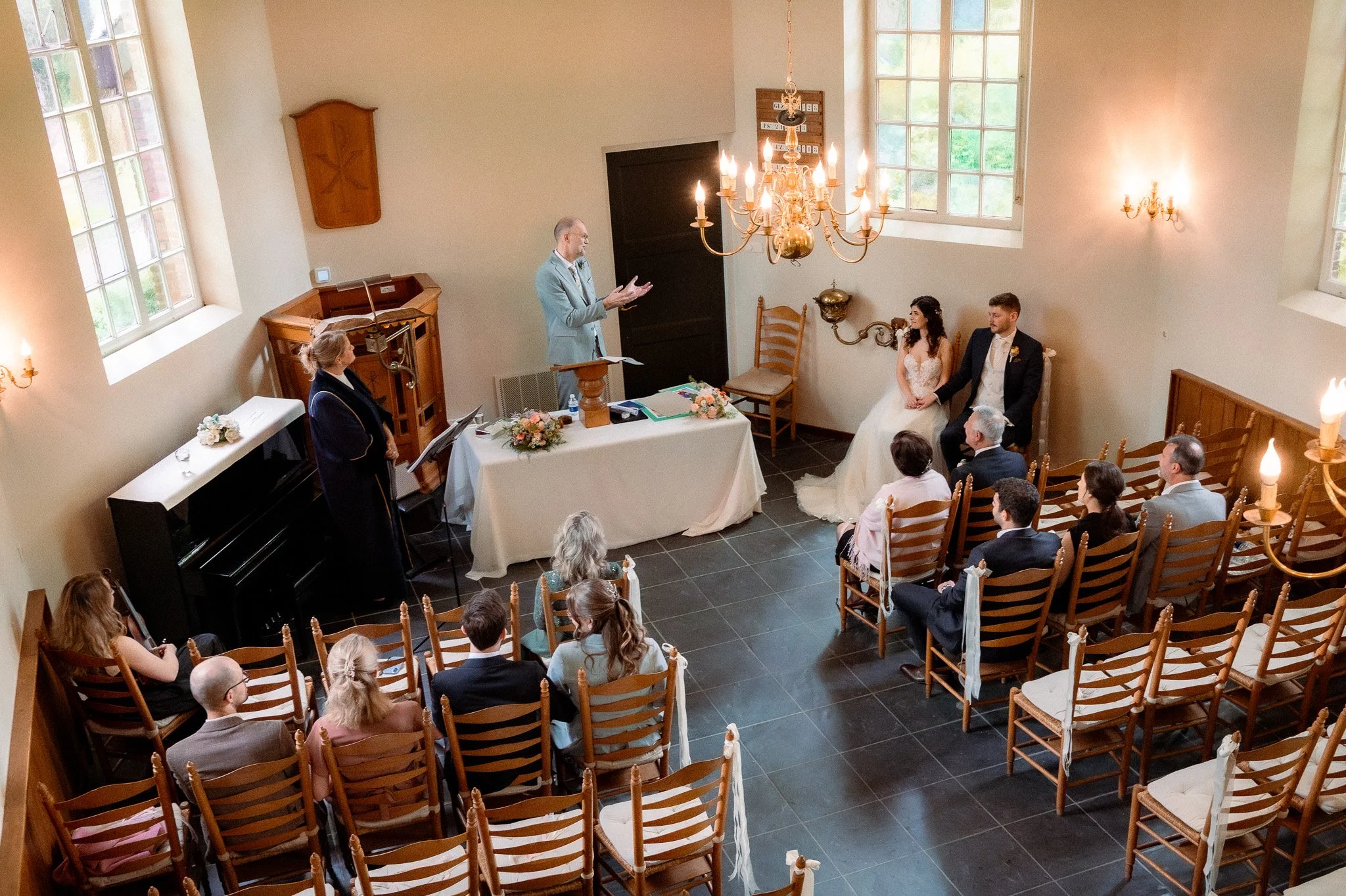 A wedding ceremony taking place in a small, warmly lit room with a high ceiling, large windows, a chandelier, and wooden chairs. The bride and groom sit together at the front, facing the officiant, with friends and family seated around them.