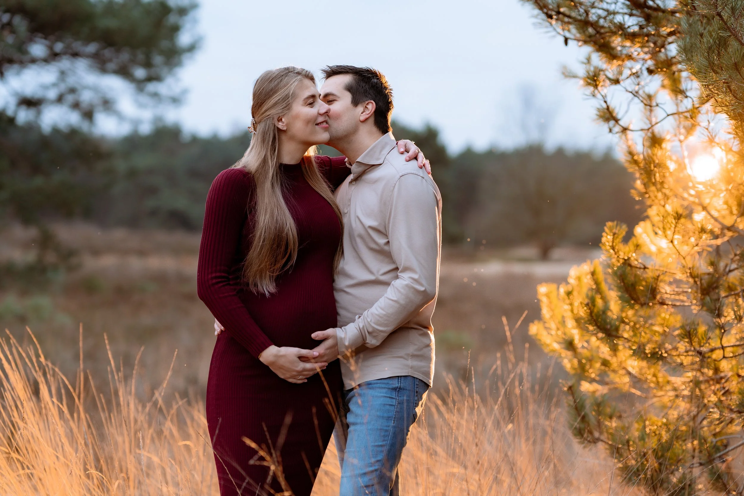 A pregnant woman and a man kissing outdoors during sunset, with the woman holding her belly, standing among tall grass and trees.