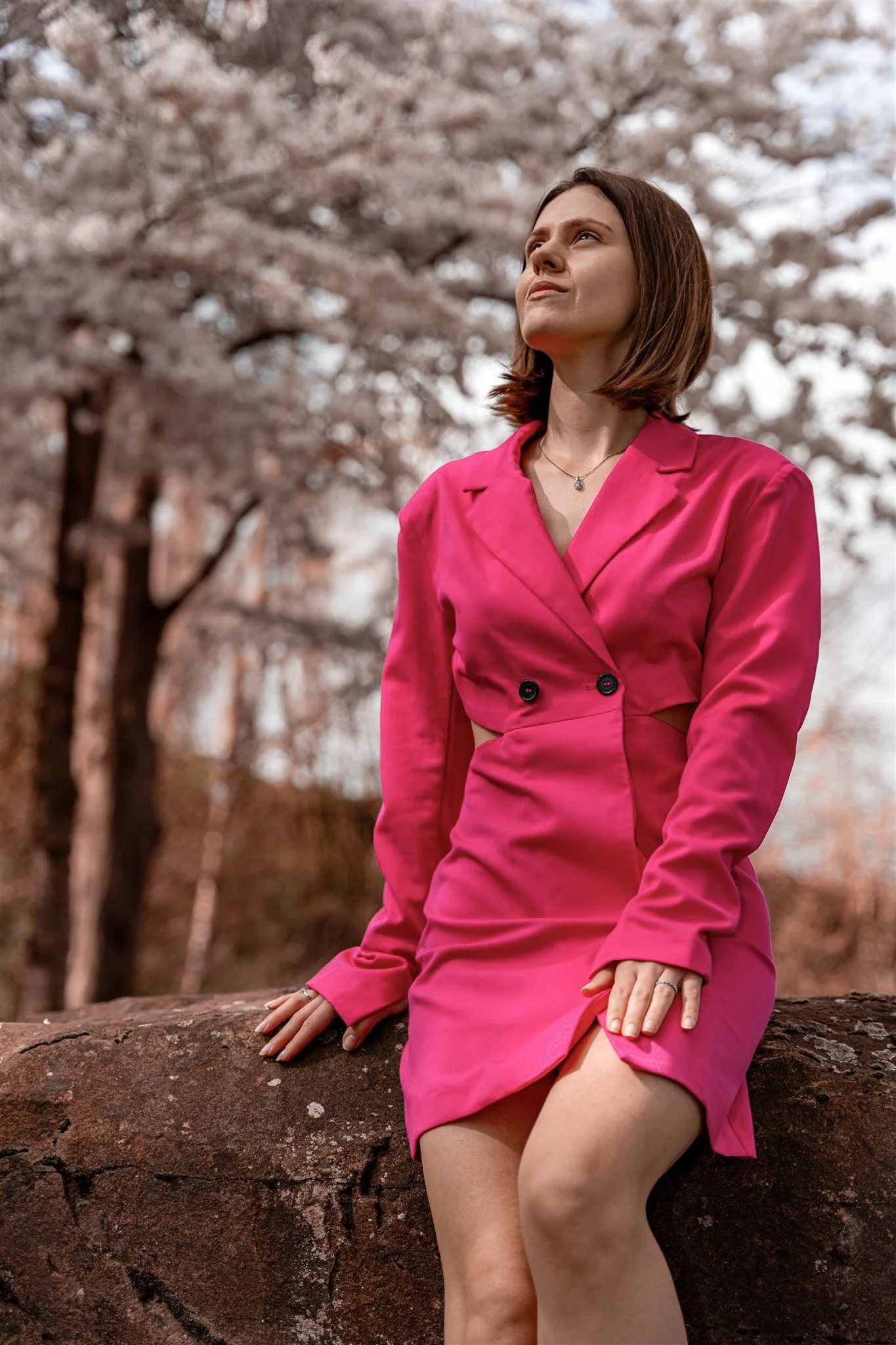 A woman in a pink dress sitting on a rock outdoors, with cherry blossoms in the background.