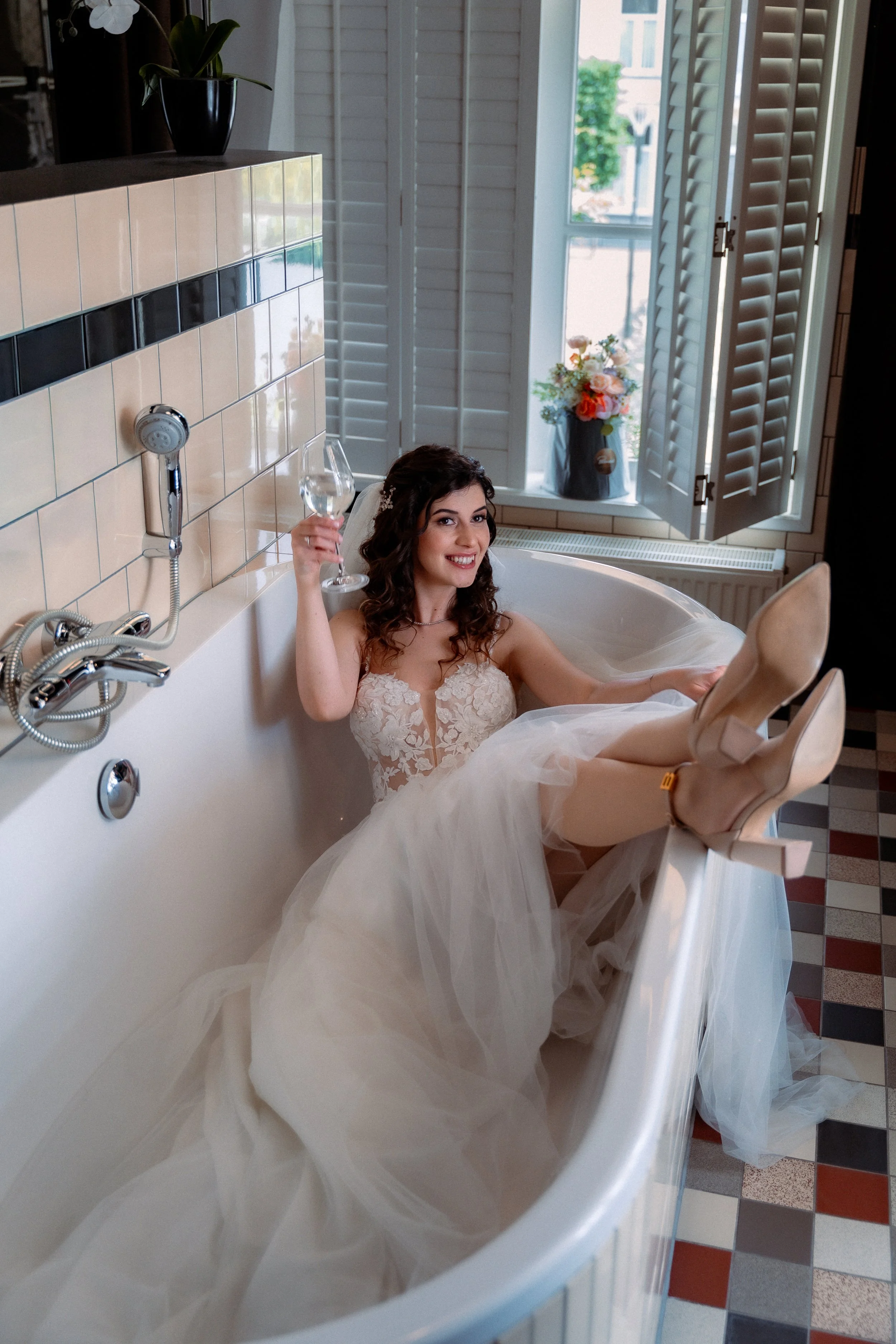 A woman in a wedding dress is relaxing in a bathtub, holding a glass of wine, with her legs raised and feet in high heels.