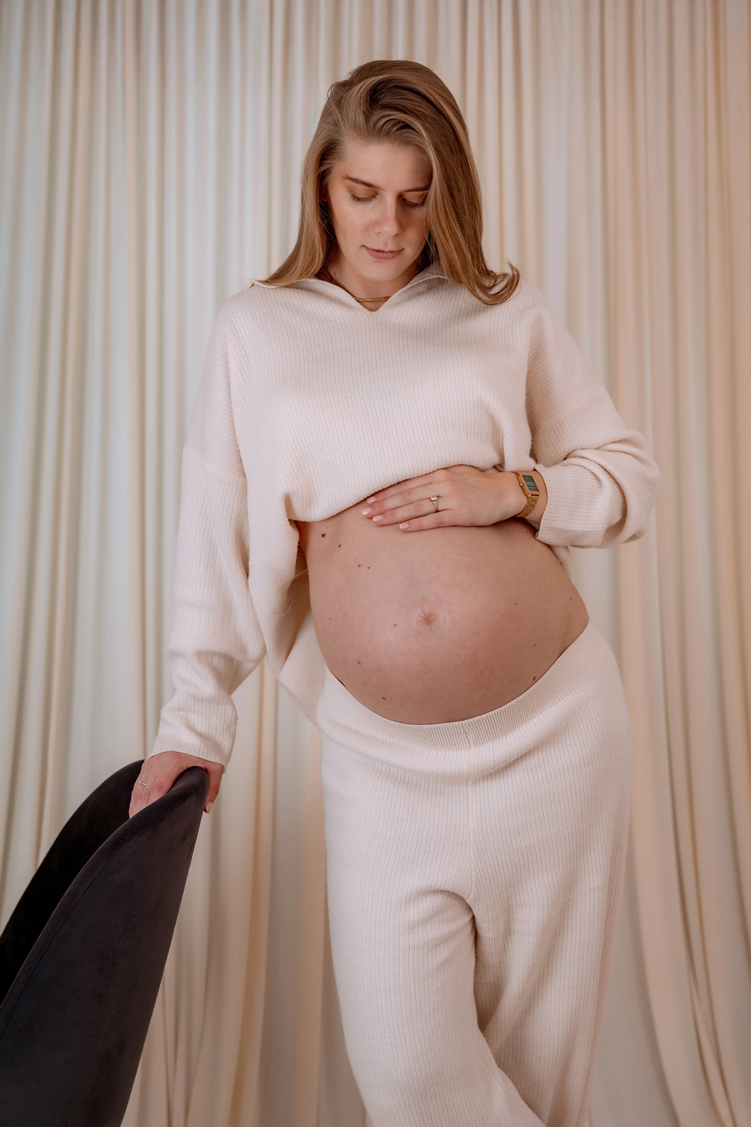 A pregnant woman in cream-colored clothing looking down and touching her belly, standing in front of cream-colored curtains.