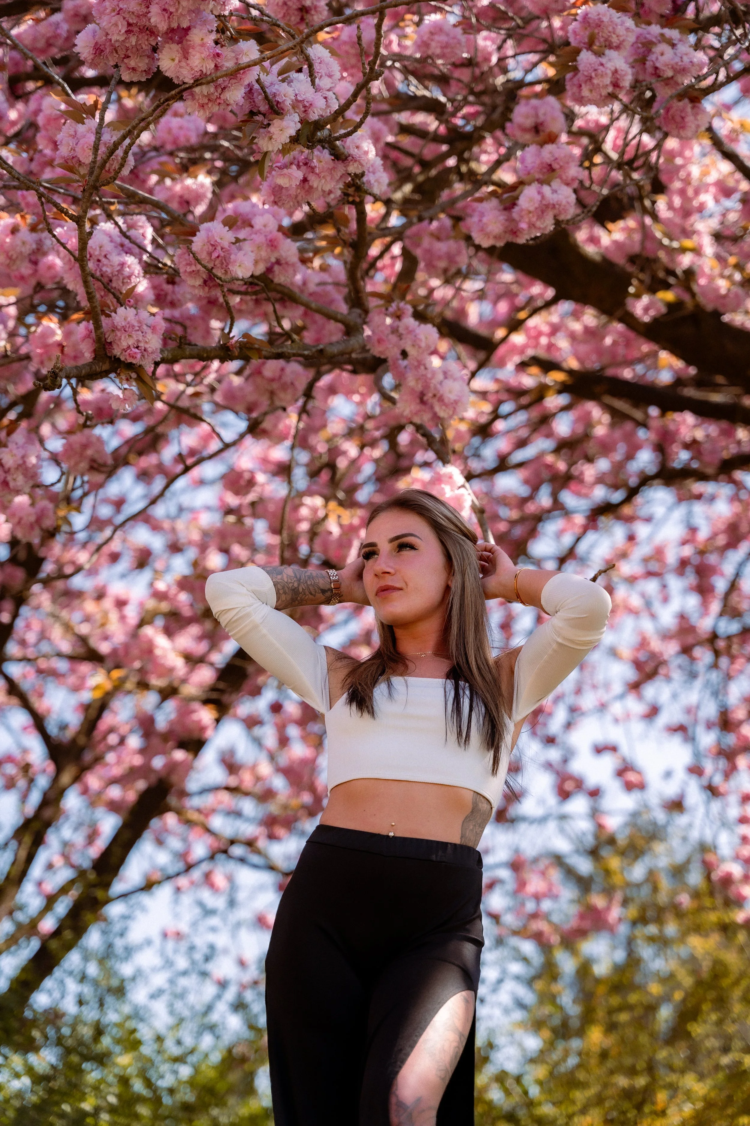 A woman standing outdoors under blooming pink cherry blossom trees, wearing a white crop top and black skirt with a thigh-high slit, with her arms raised behind her head.