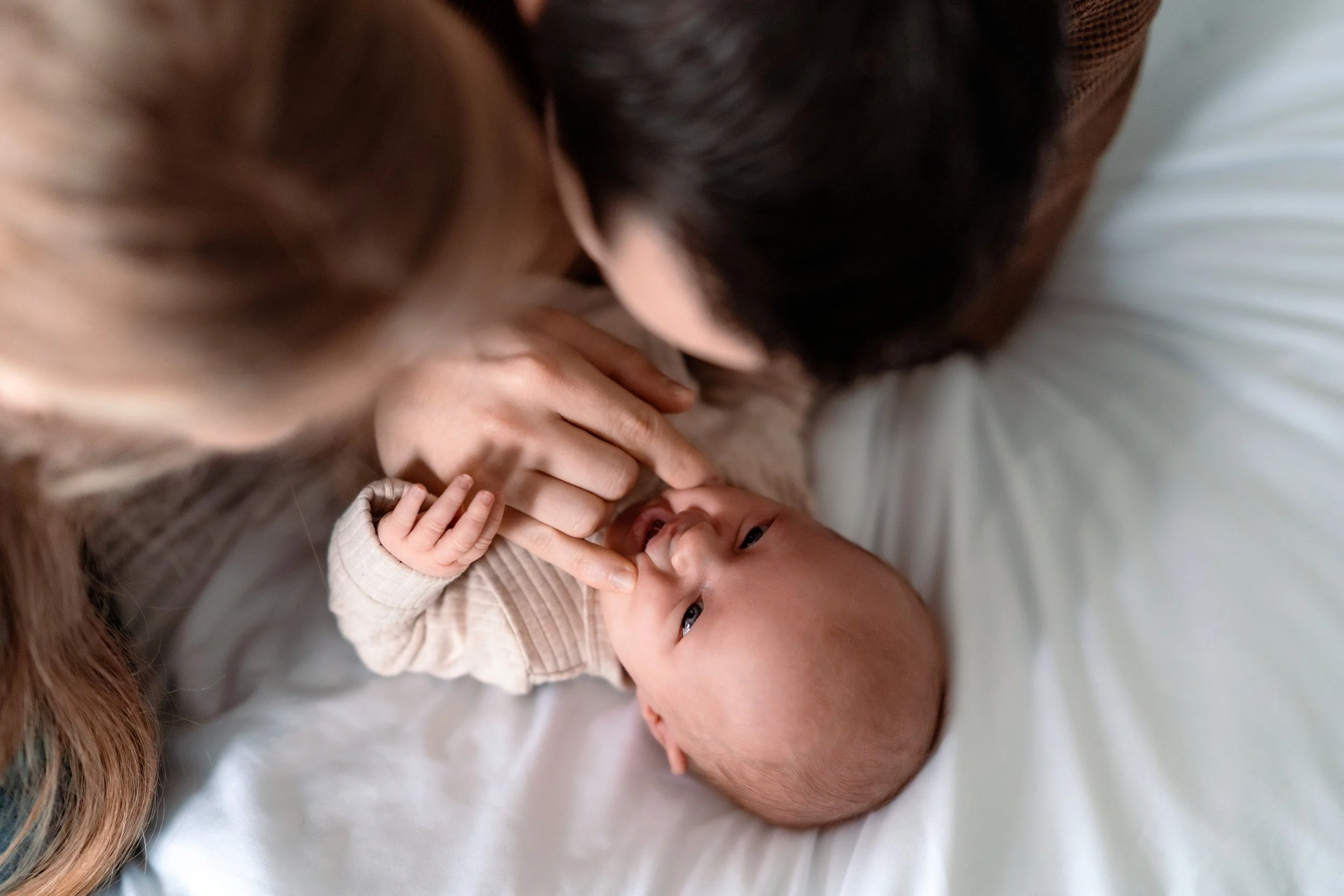 A baby lying on a white bed looking up, with a woman and a man leaning over and touching the baby's nose.