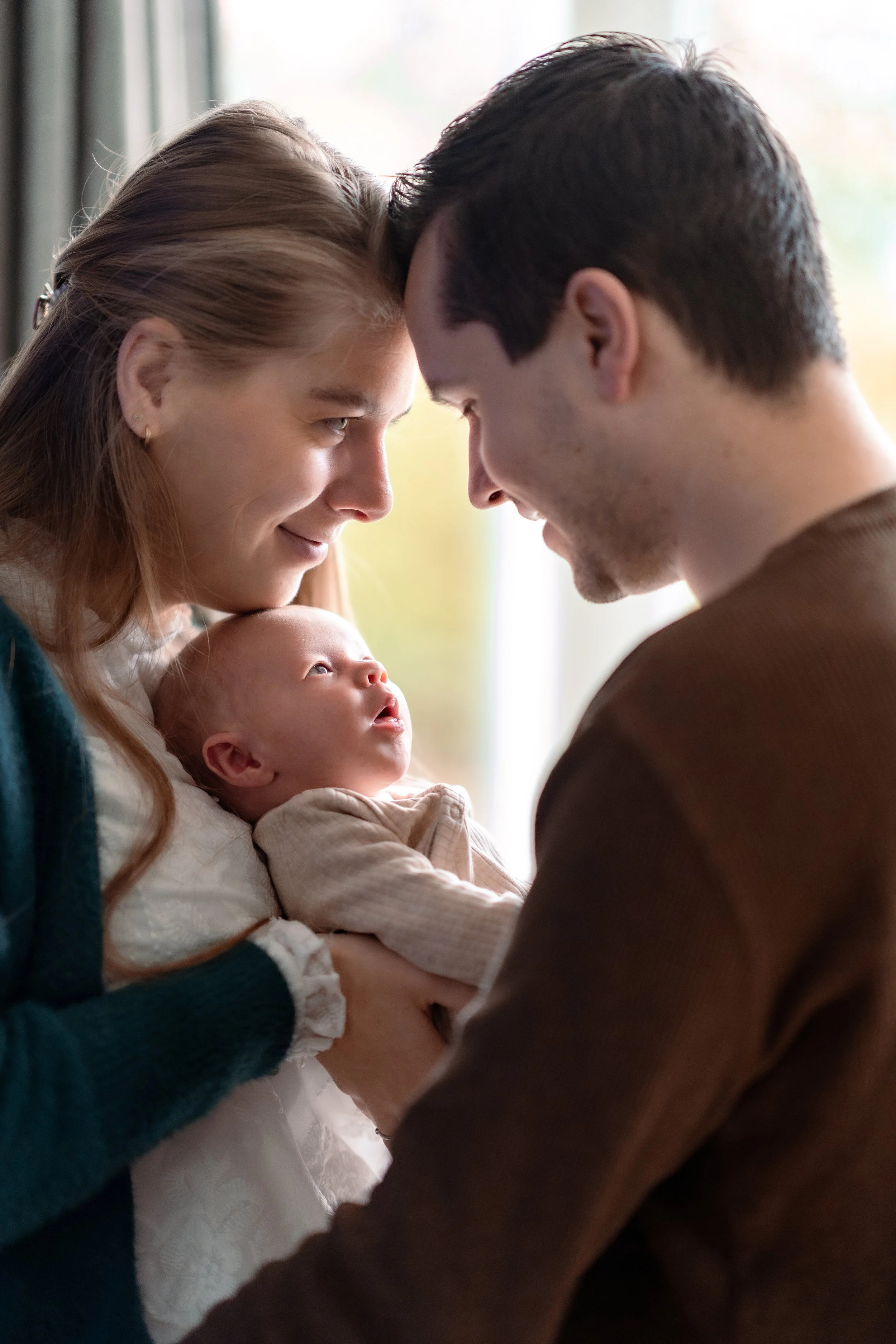 A young family with a mother, father, and infant child looking at each other lovingly in a cozy indoor setting.