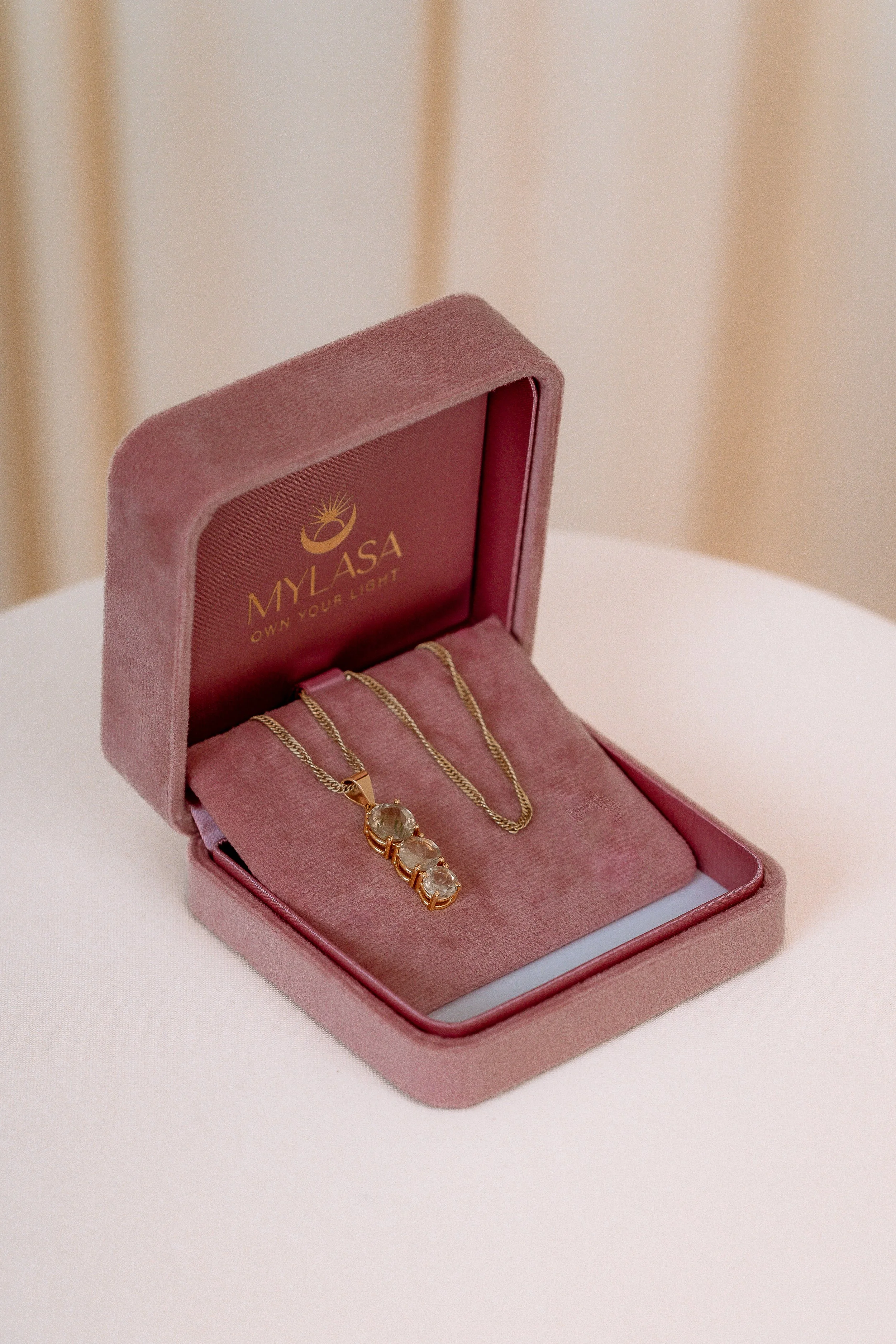 A pink jewelry box with gold velvet interior containing a pendant necklace and a chain, with the brand name 'MYLASA' and the slogan 'Own Your Light' inside the lid.