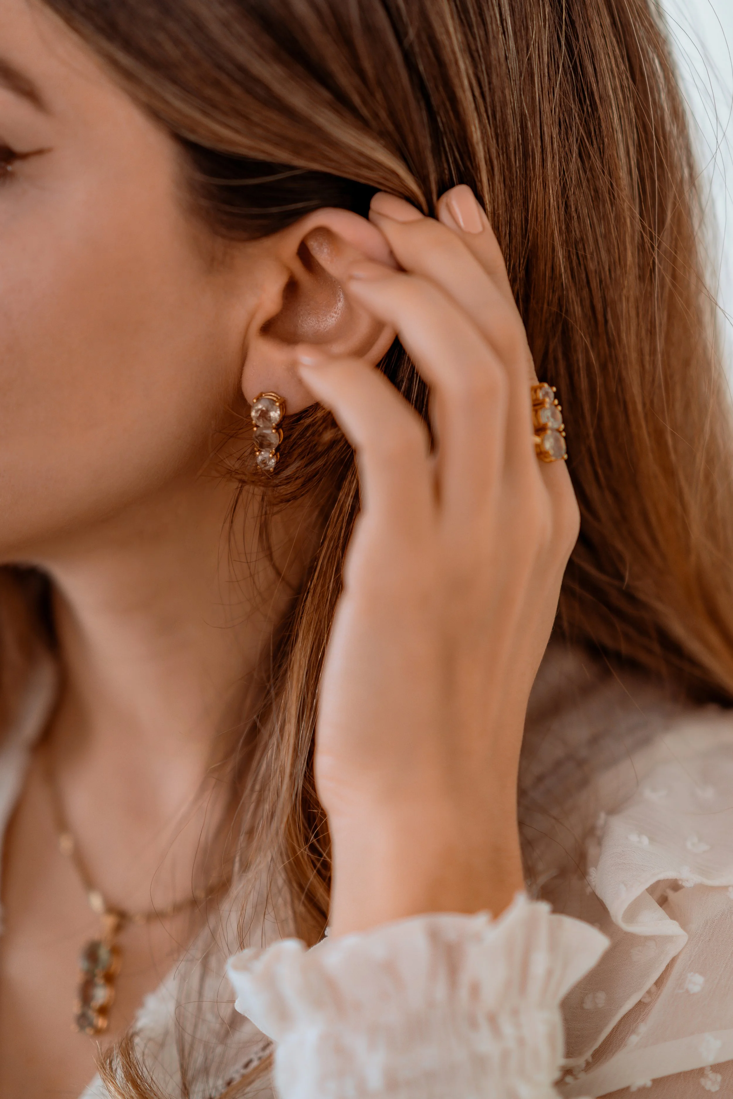 Close-up of a woman with auburn hair adjusting an earring with her hand, wearing jewelry including a necklace and a ring, dressed in a white outfit with ruffled sleeves.