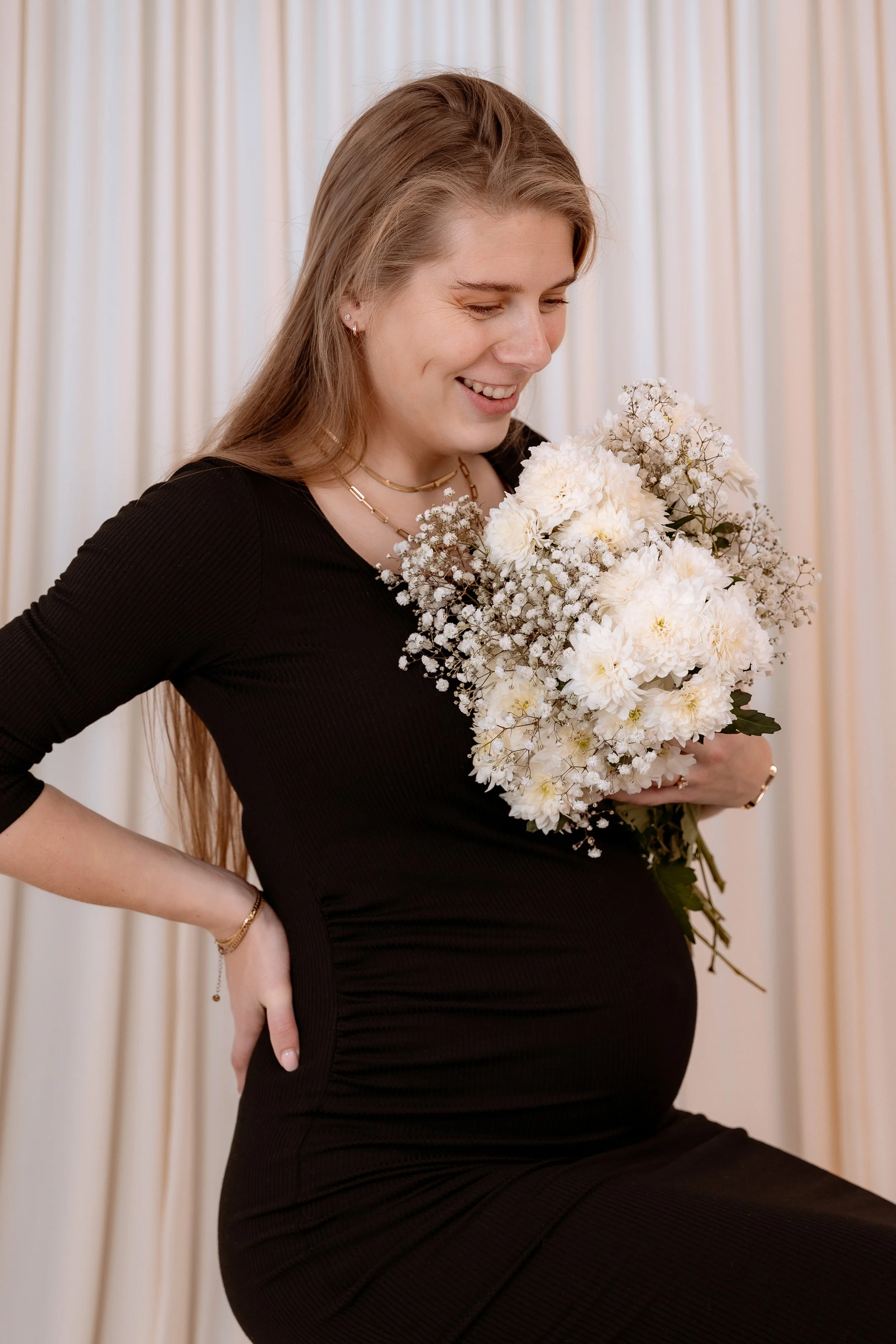 A pregnant woman in a black dress holding a bouquet of white flowers, smiling, with a curtain background.