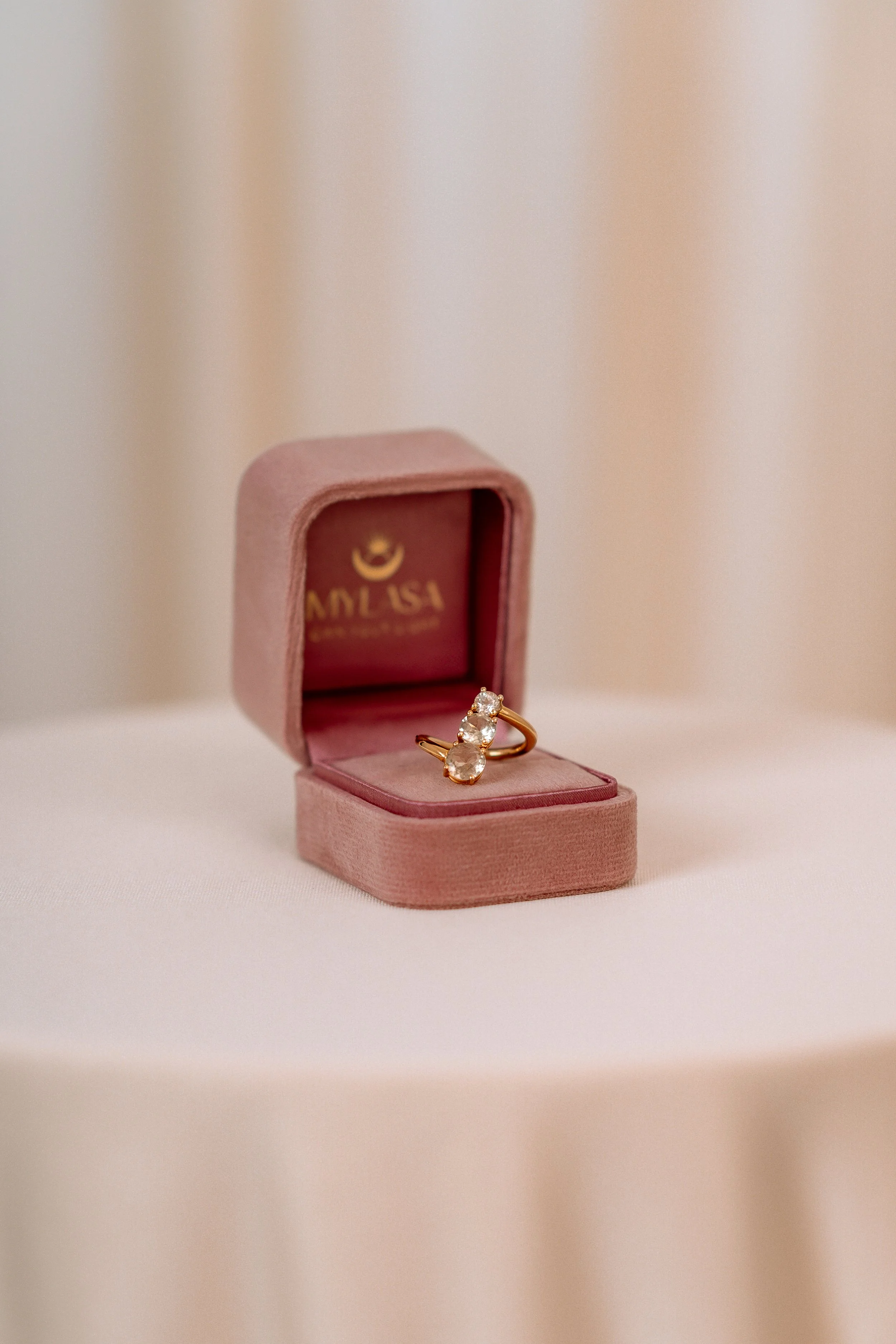 A gold ring with three clear gemstones placed inside a pink velvet jewelry box on a white surface.