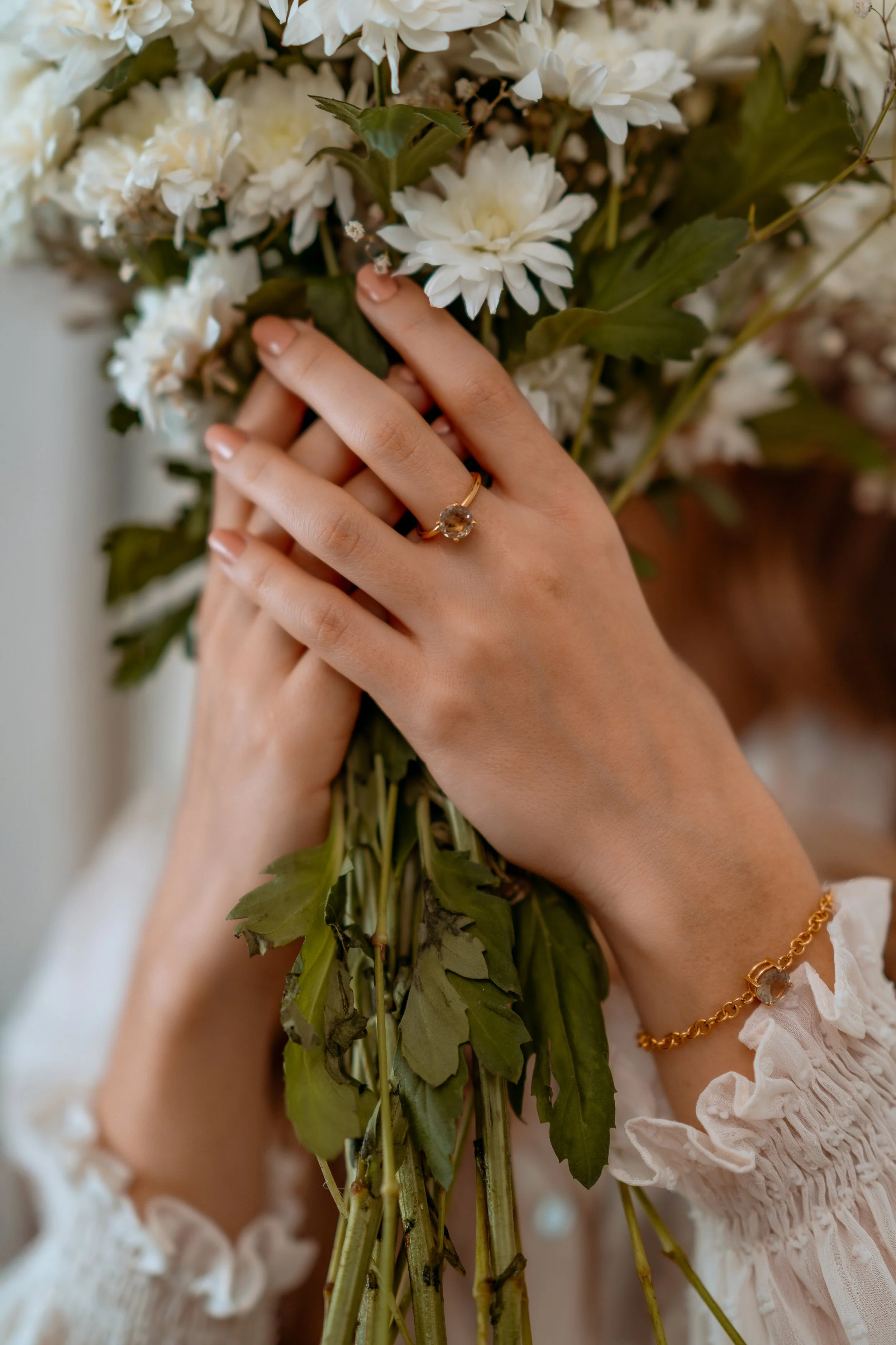 Close-up of a woman's hands holding a bouquet of white flowers, wearing a gold ring with a large gemstone and a gold bracelet, with a white ruffled sleeve visible.