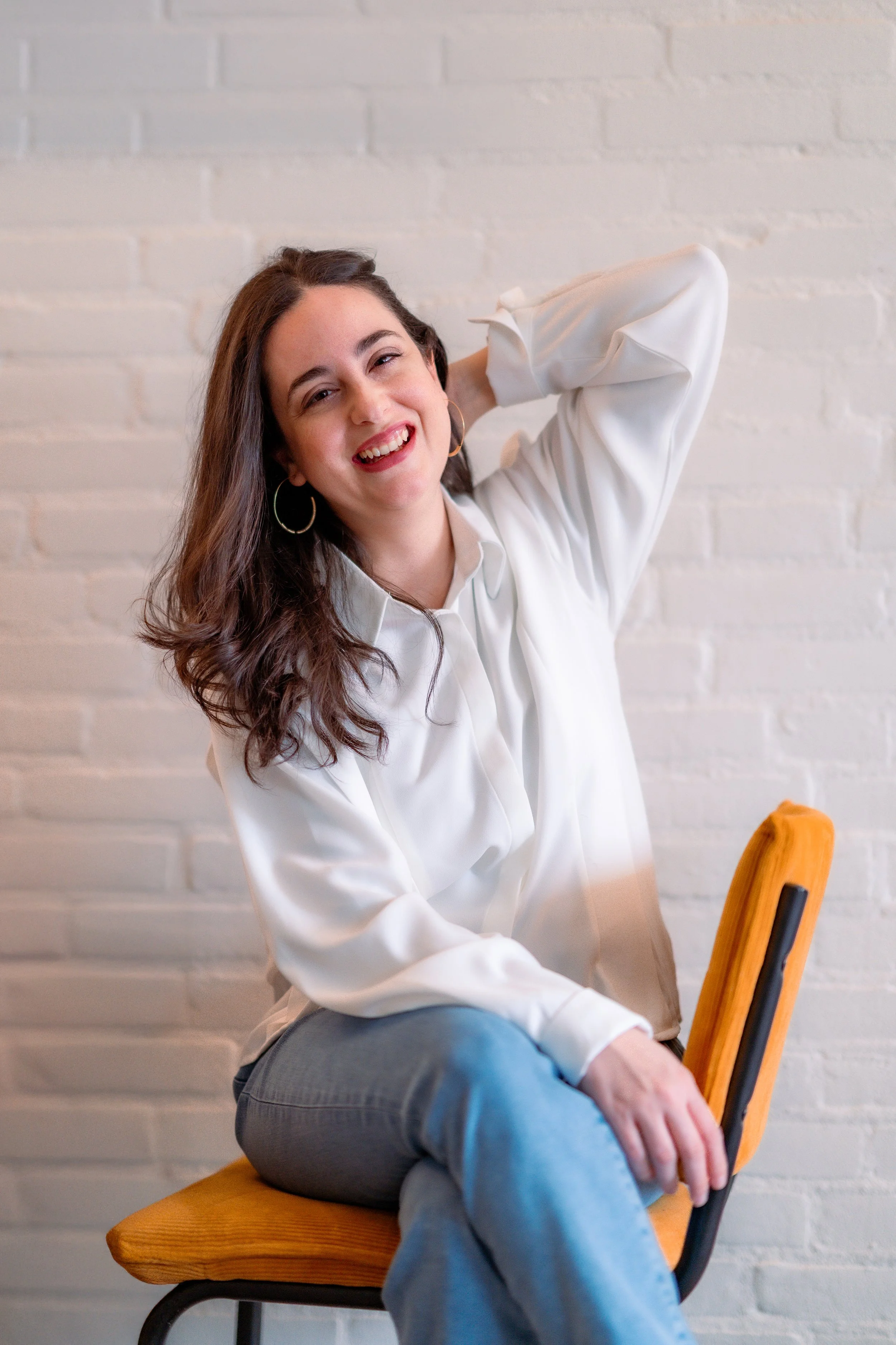A woman with long brown hair smiling, wearing a white blouse and light blue jeans, sitting on a yellow chair with a white brick wall in the background.