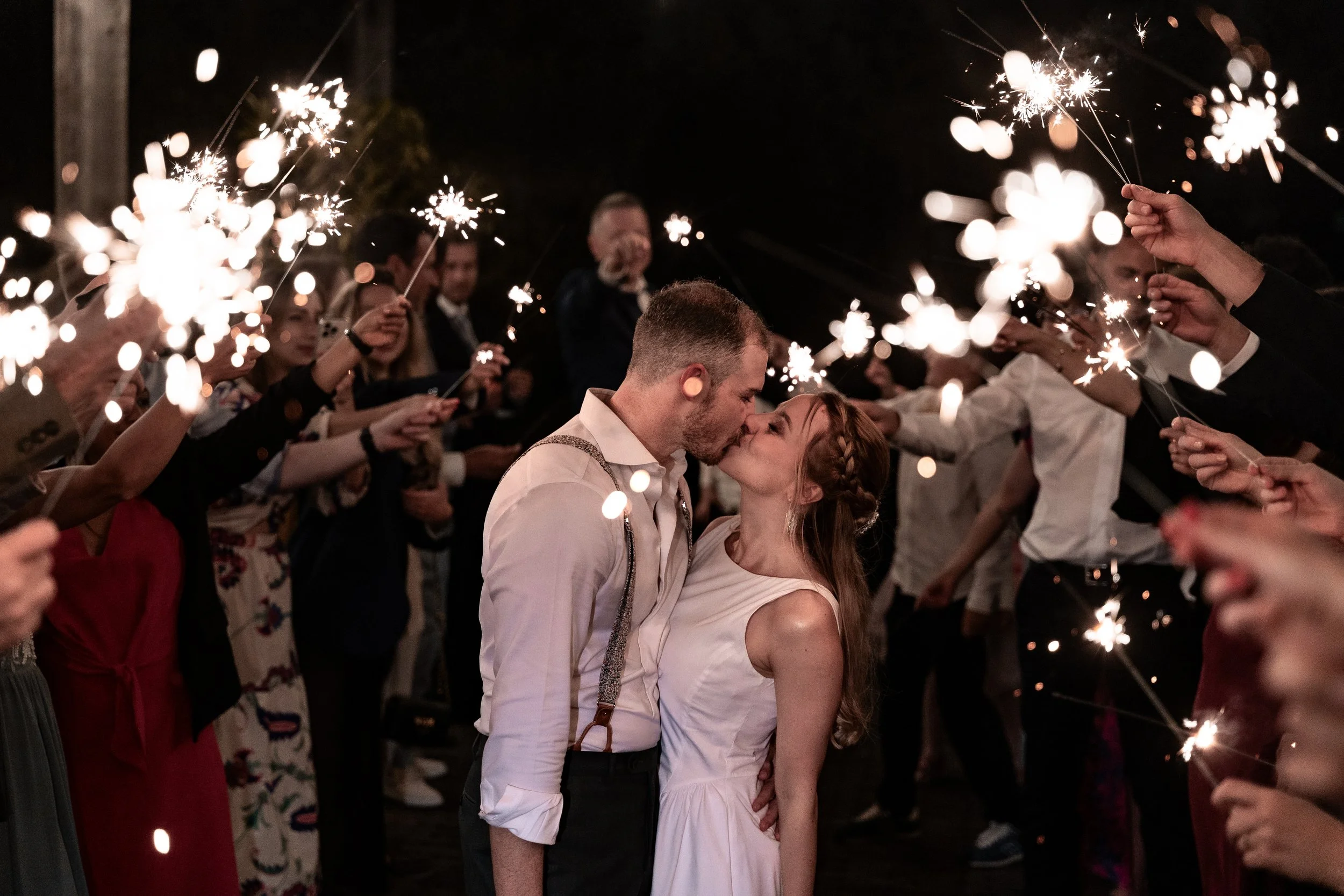 A couple kisses at a celebration with sparklers held by many people around them at night.