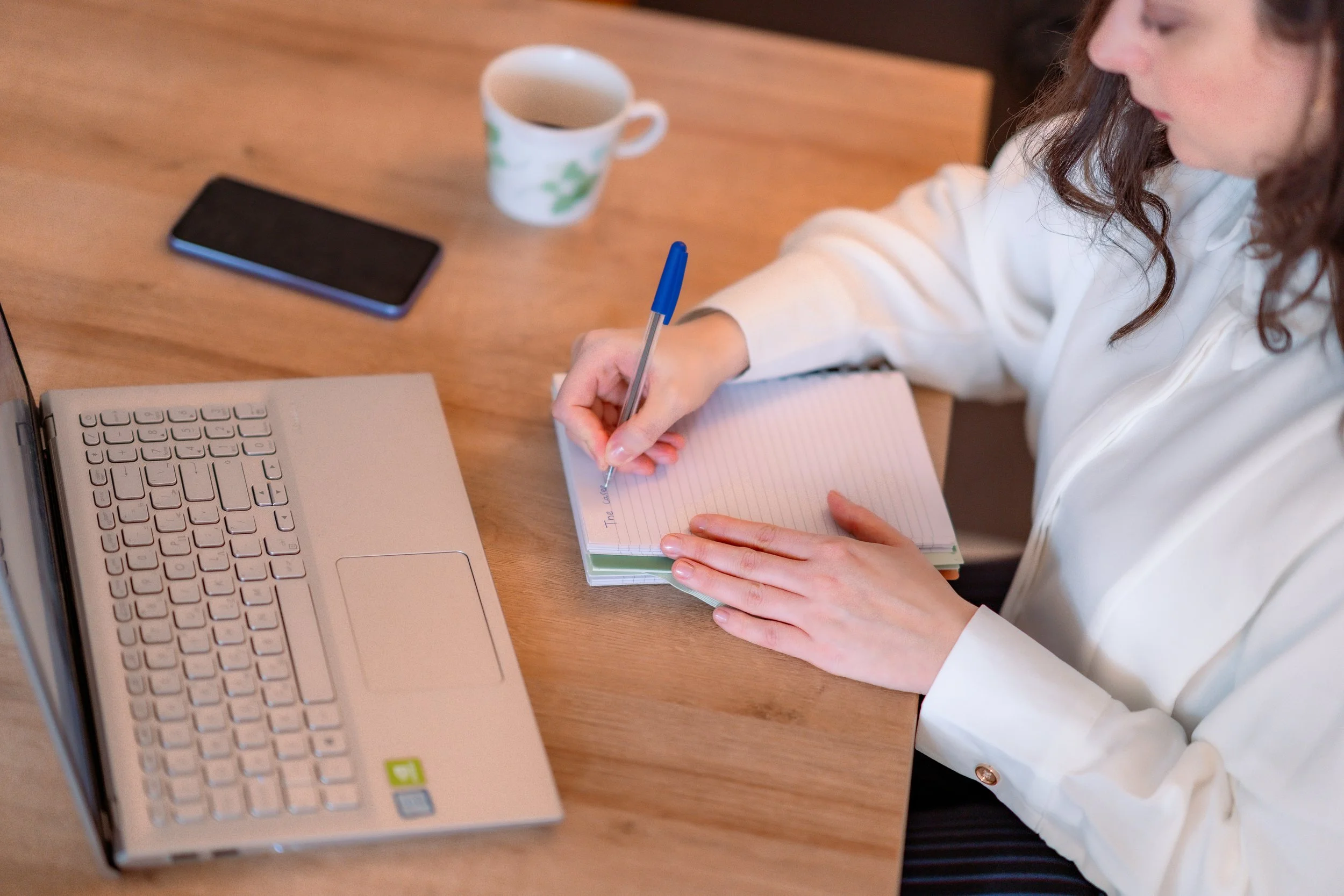A woman sitting at a wooden table, writing in a lined notebook with a blue pen, with a laptop, a smartphone, a mug, and a white sweater partially visible.