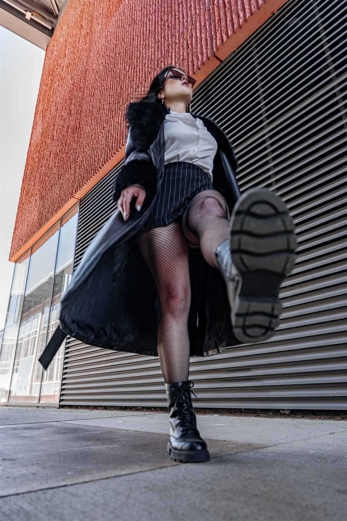 A woman dressed in a black coat, pinstripe skirt, fishnet stockings, and combat boots is walking with her leg raised high, capturing a low-angle perspective outdoors near a modern building with orange and gray exterior walls.