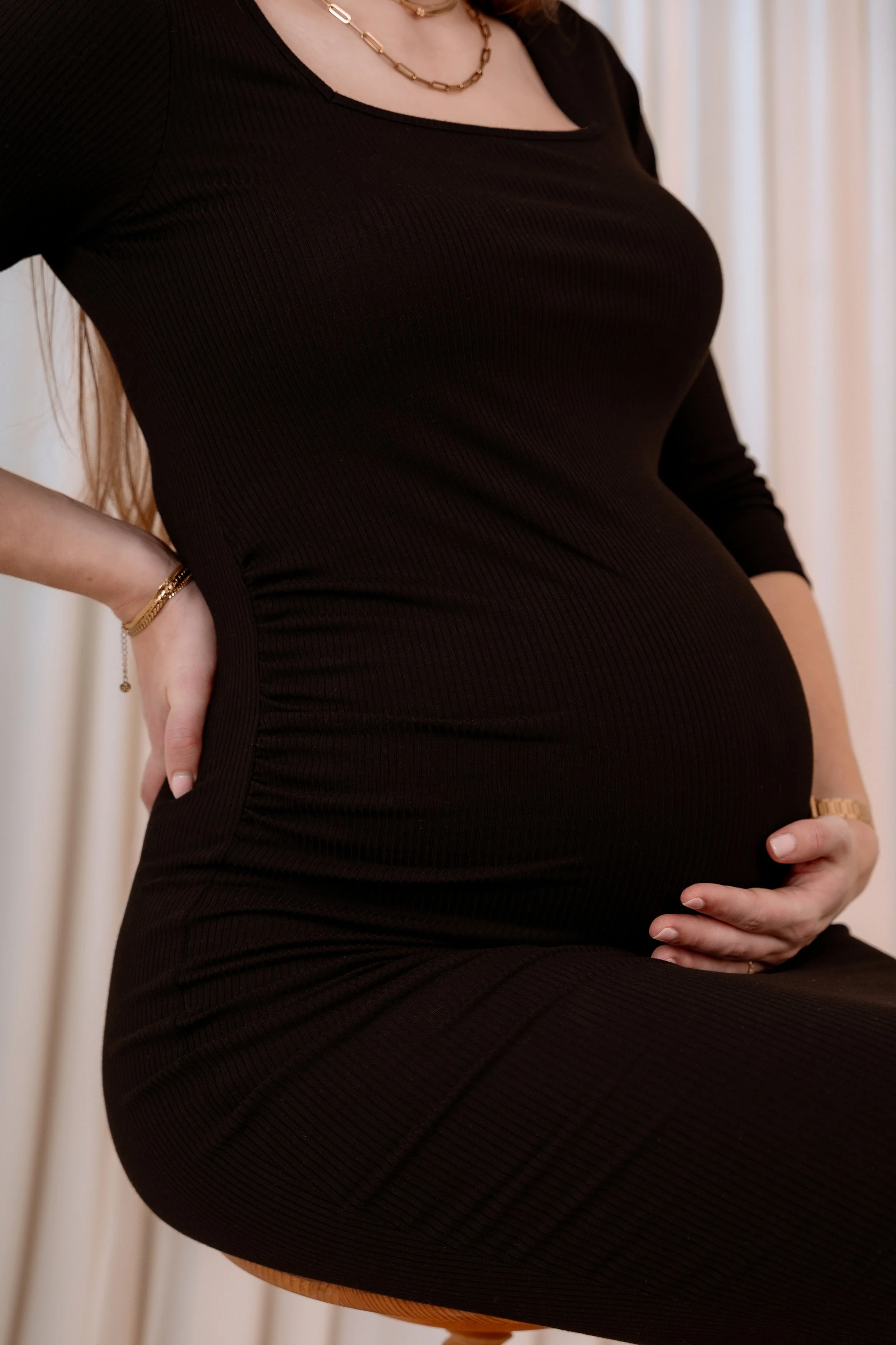 A pregnant woman wearing a black dress, gold necklace, bracelet, and ring, with her hand resting on her belly.