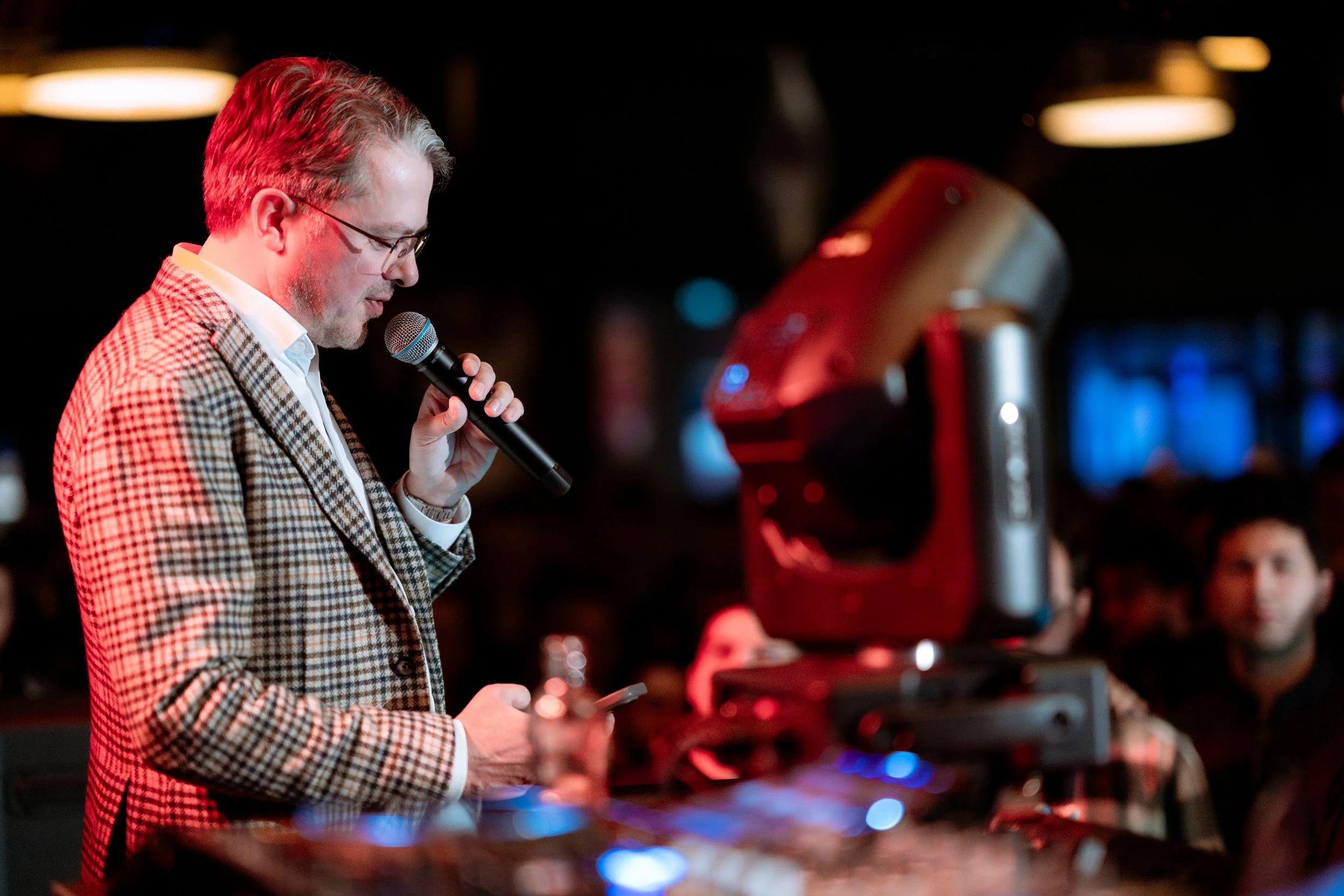 A man with glasses and a checkered blazer speaking into a microphone at an indoor event, with audience members in the background and a large speaker nearby.