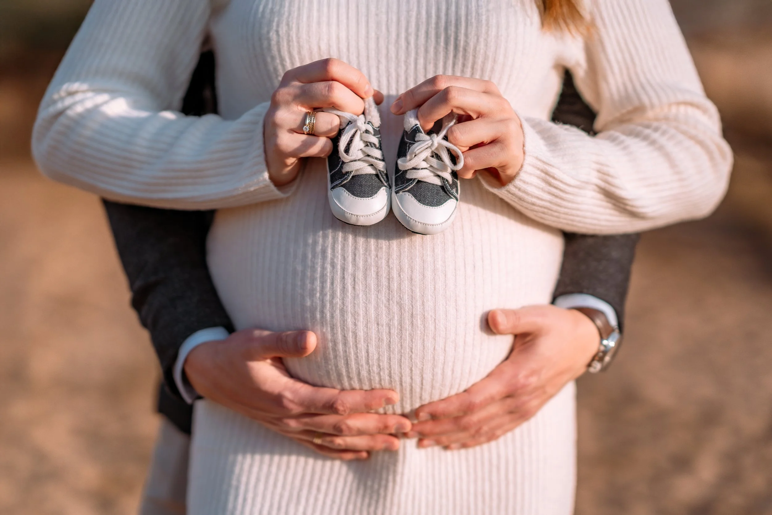 A pregnant woman holding a pair of miniature sneakers above her belly, with another person helping to support her, outdoors in natural light.