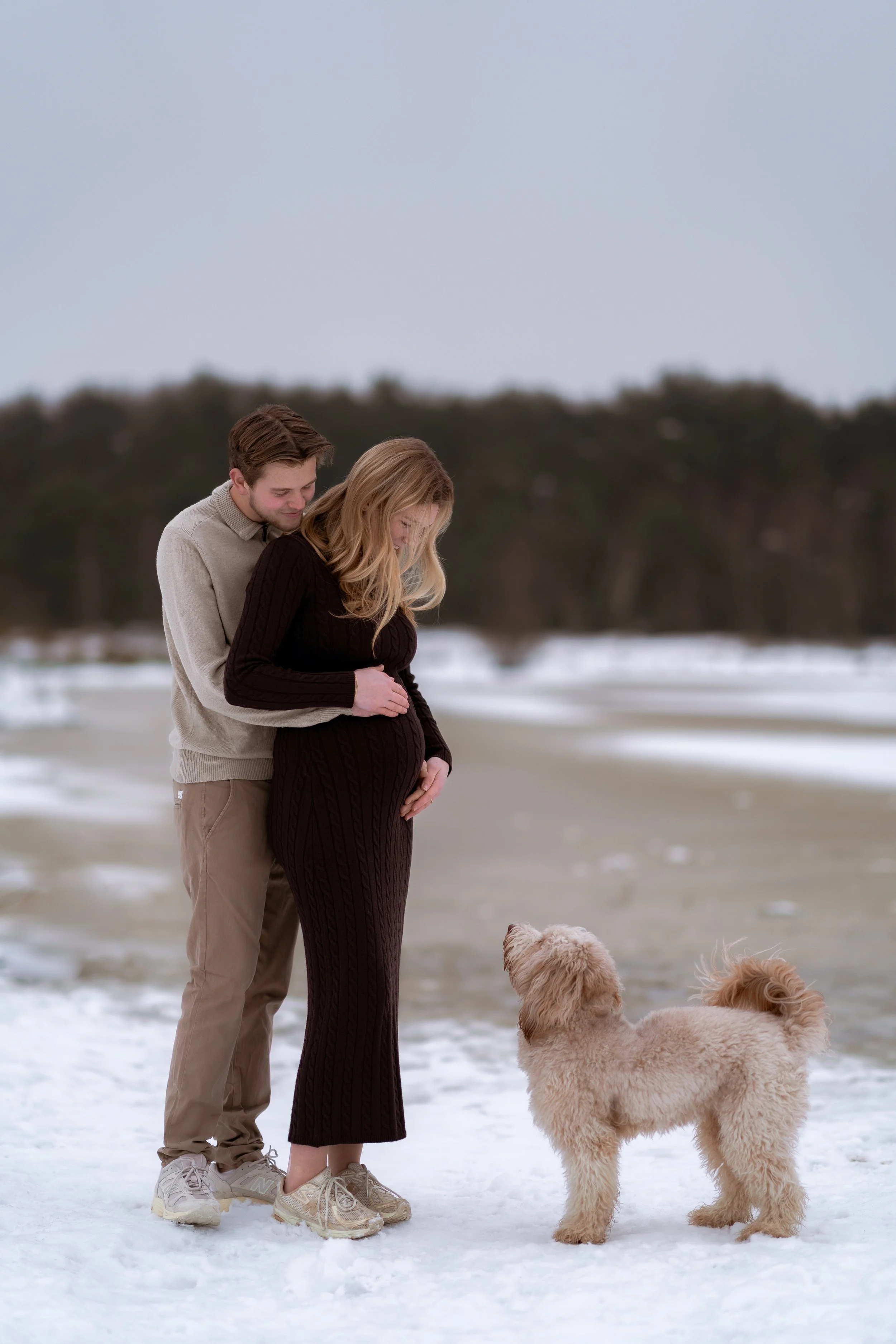 A pregnant woman and a man standing on snow-covered ground at the beach, looking down at a fluffy dog.
