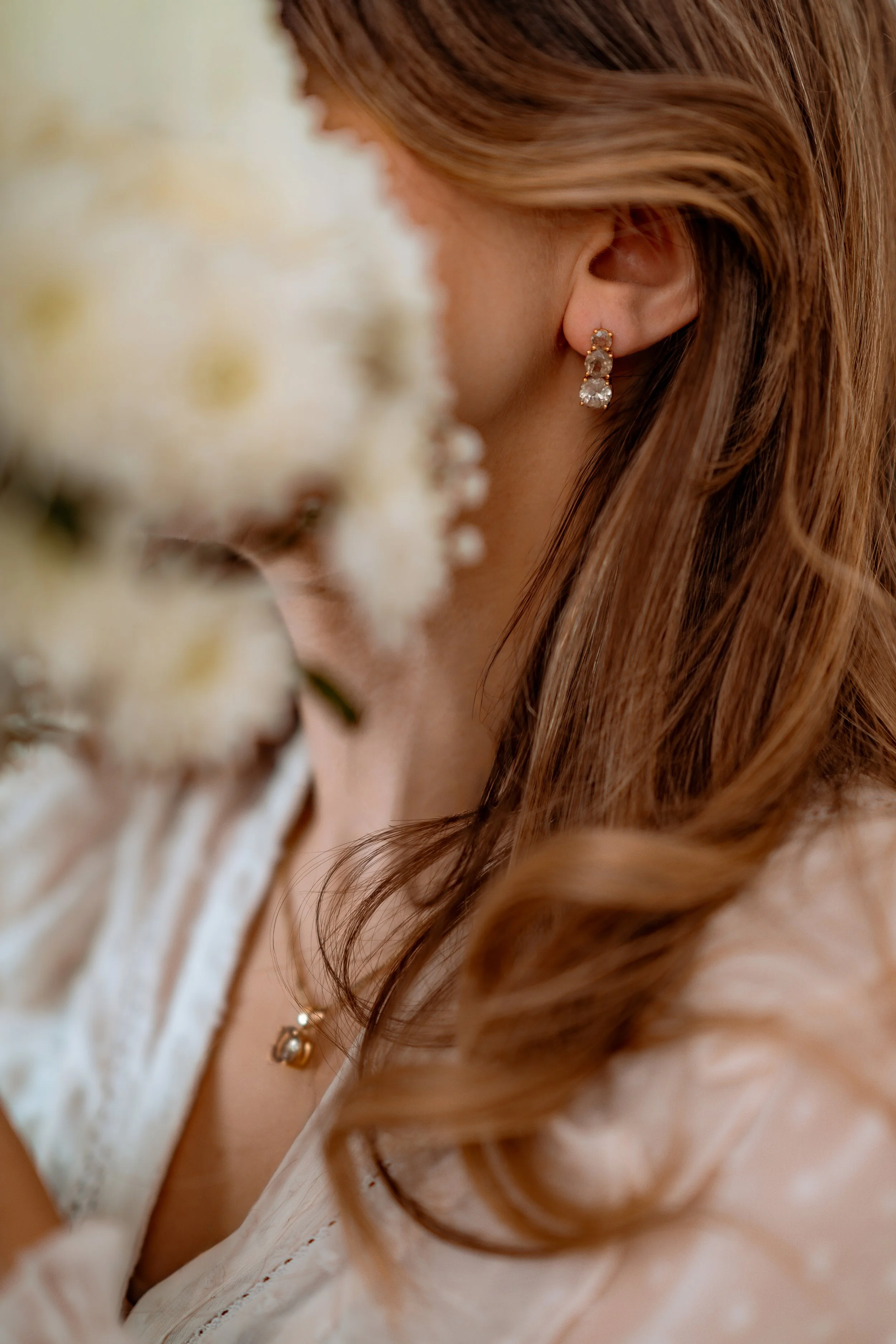 Close-up of a woman's ear with diamond drop earrings, surrounded by brown hair and white flowers, blurred in the foreground.