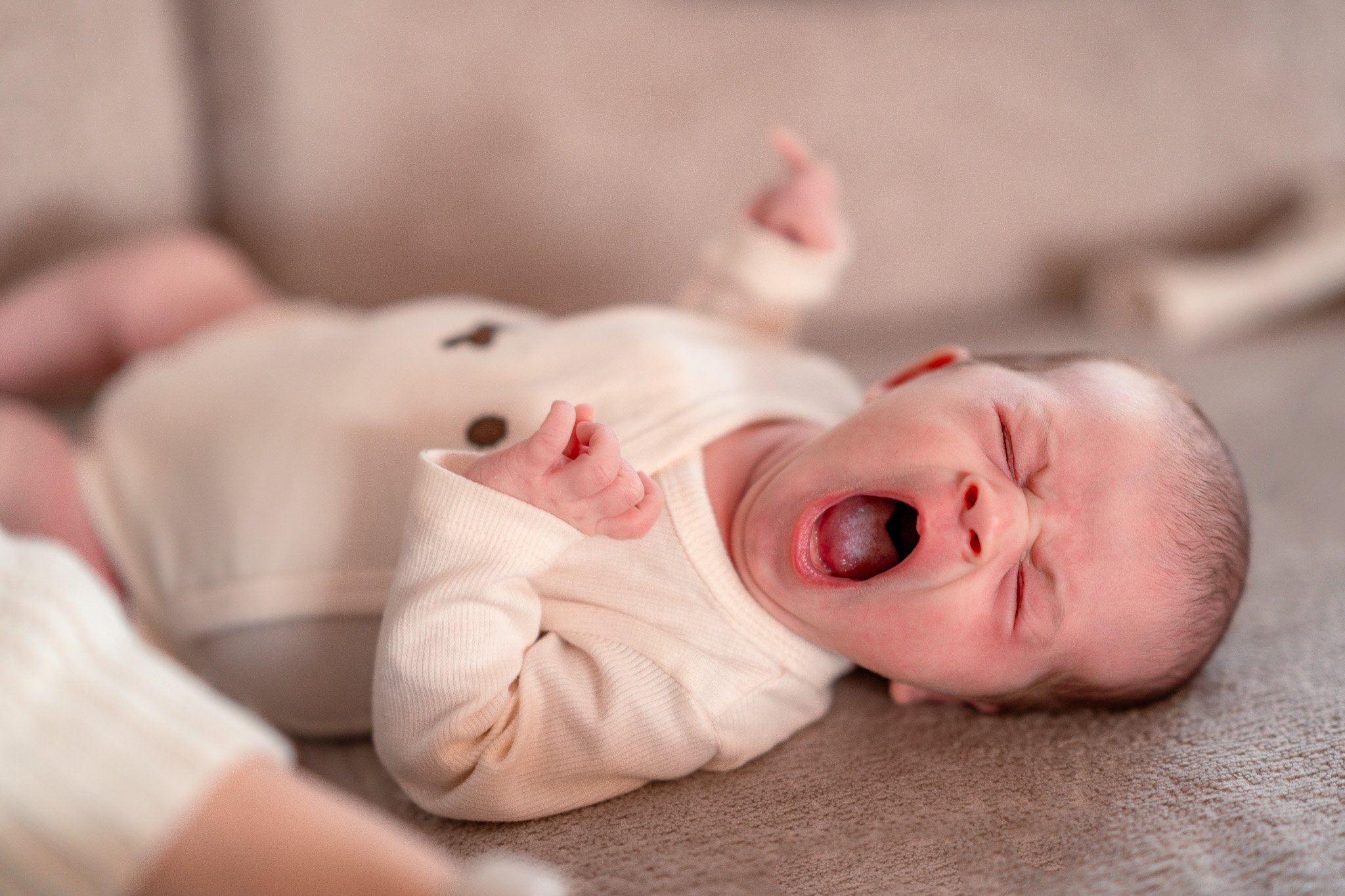 A newborn baby lying on a beige surface, yawning with eyes closed, wearing a light-colored onesie with dark buttons.