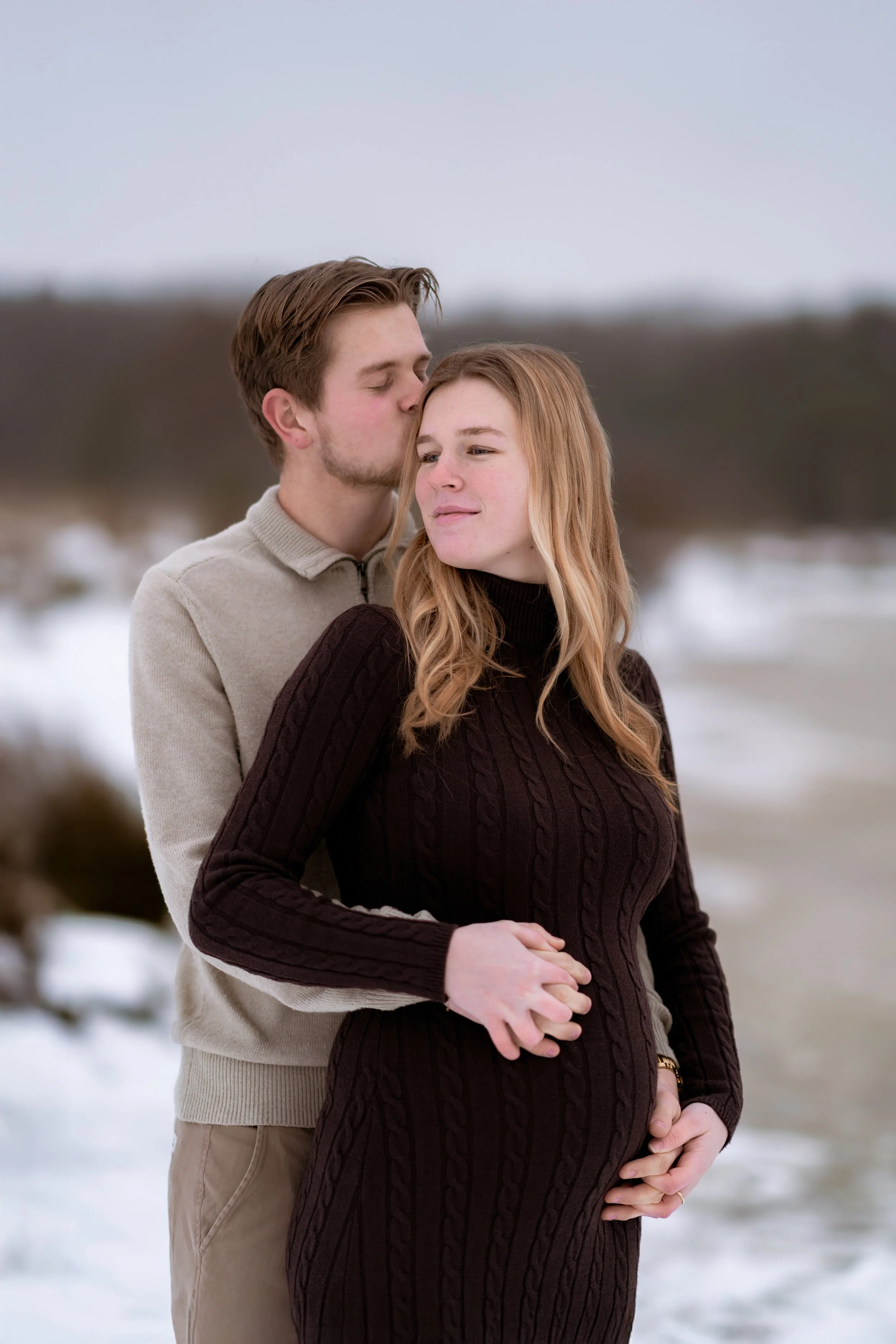 A couple standing outdoors in winter. The man is kissing the woman on the forehead, and she is smiling softly while looking away. The woman is pregnant, and her hand rests on her belly. Snow is visible on the ground in the background.