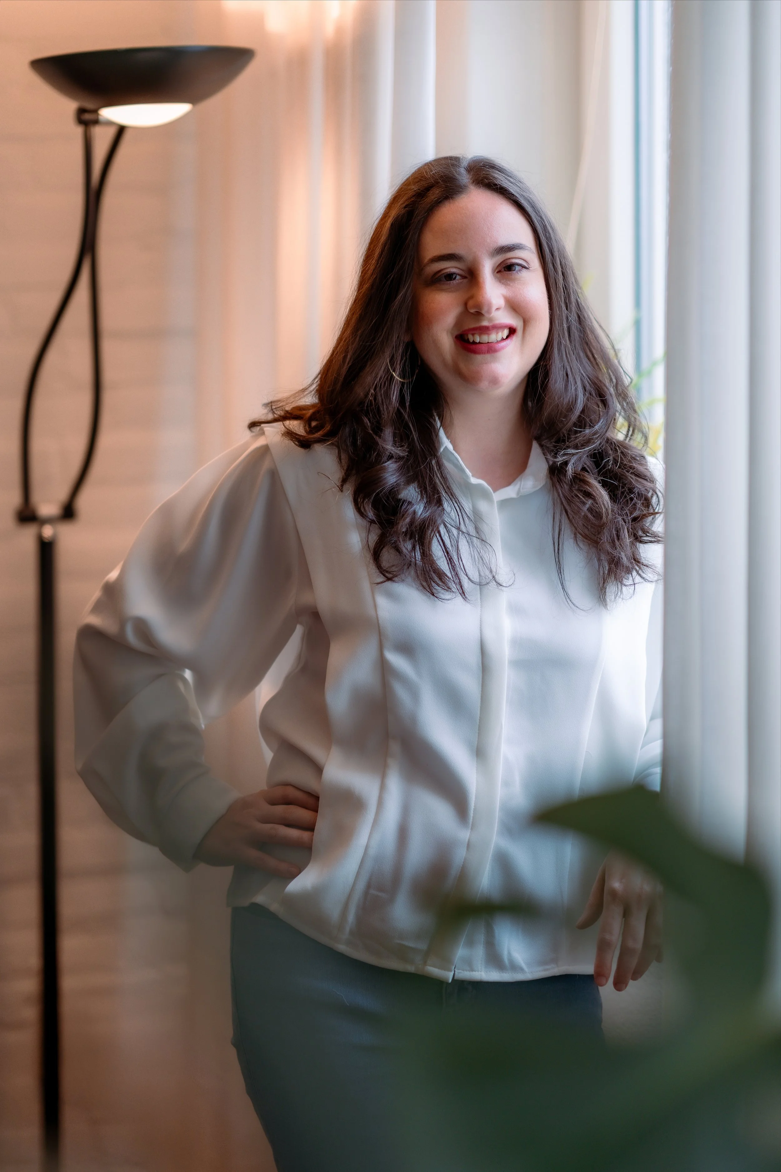 A woman with dark wavy hair wearing a white blouse, smiling and standing near a window with curtains in a well-lit room.