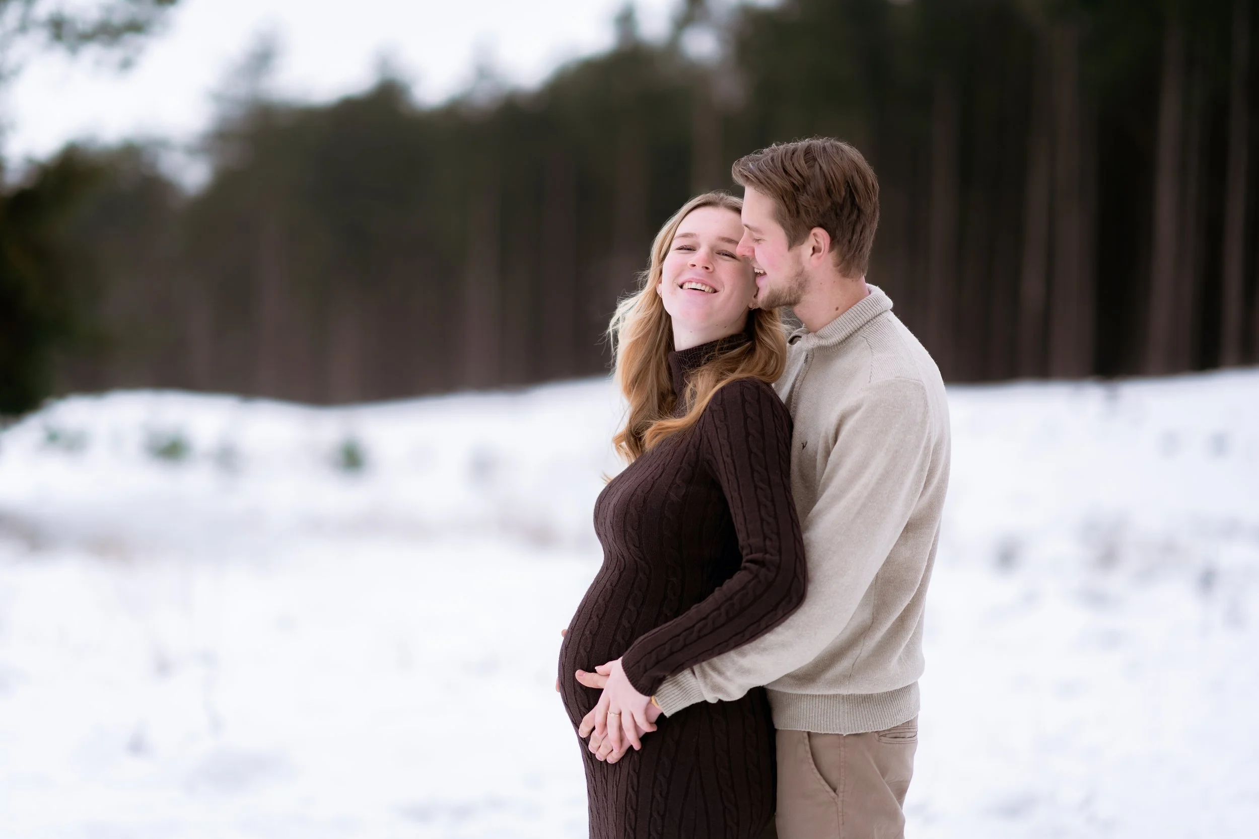 A pregnant woman and a man in an embrace outdoors in a snowy landscape surrounded by trees, smiling and enjoying each other's company.