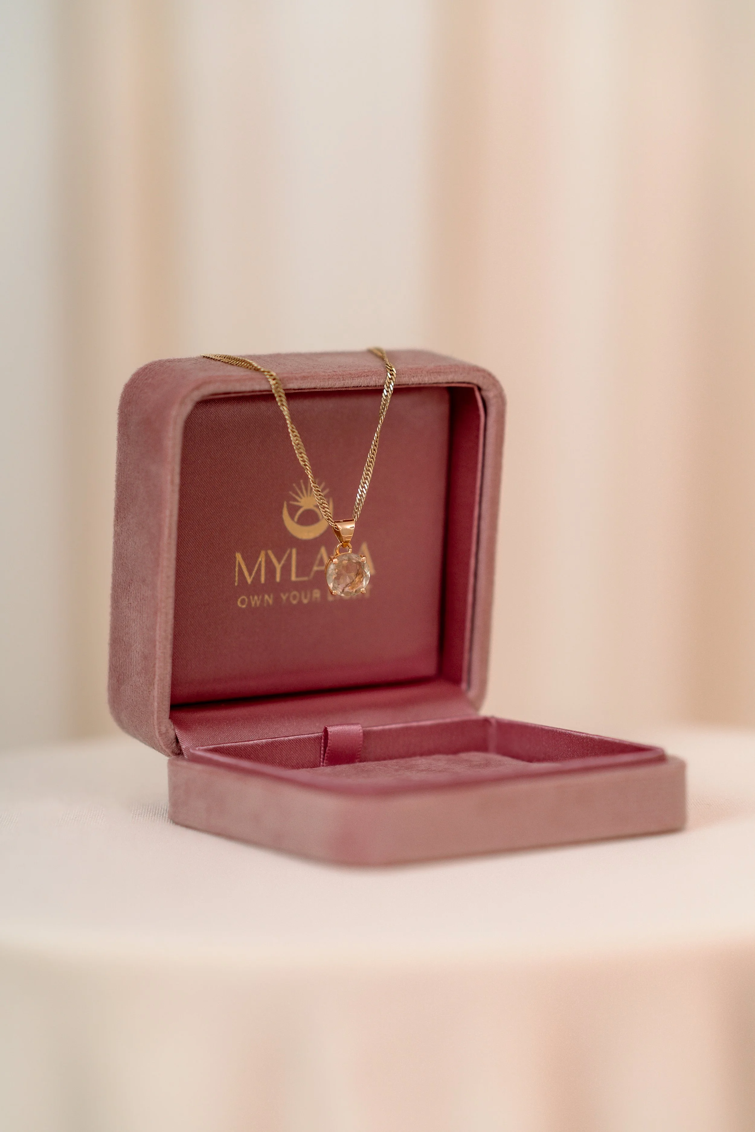 A gold necklace with a large pendant featuring a clear gemstone, displayed in an open pink jewelry box with gold interior branding that reads "MYLAQ OWN YOUR". The box is placed on a white surface against a neutral background.