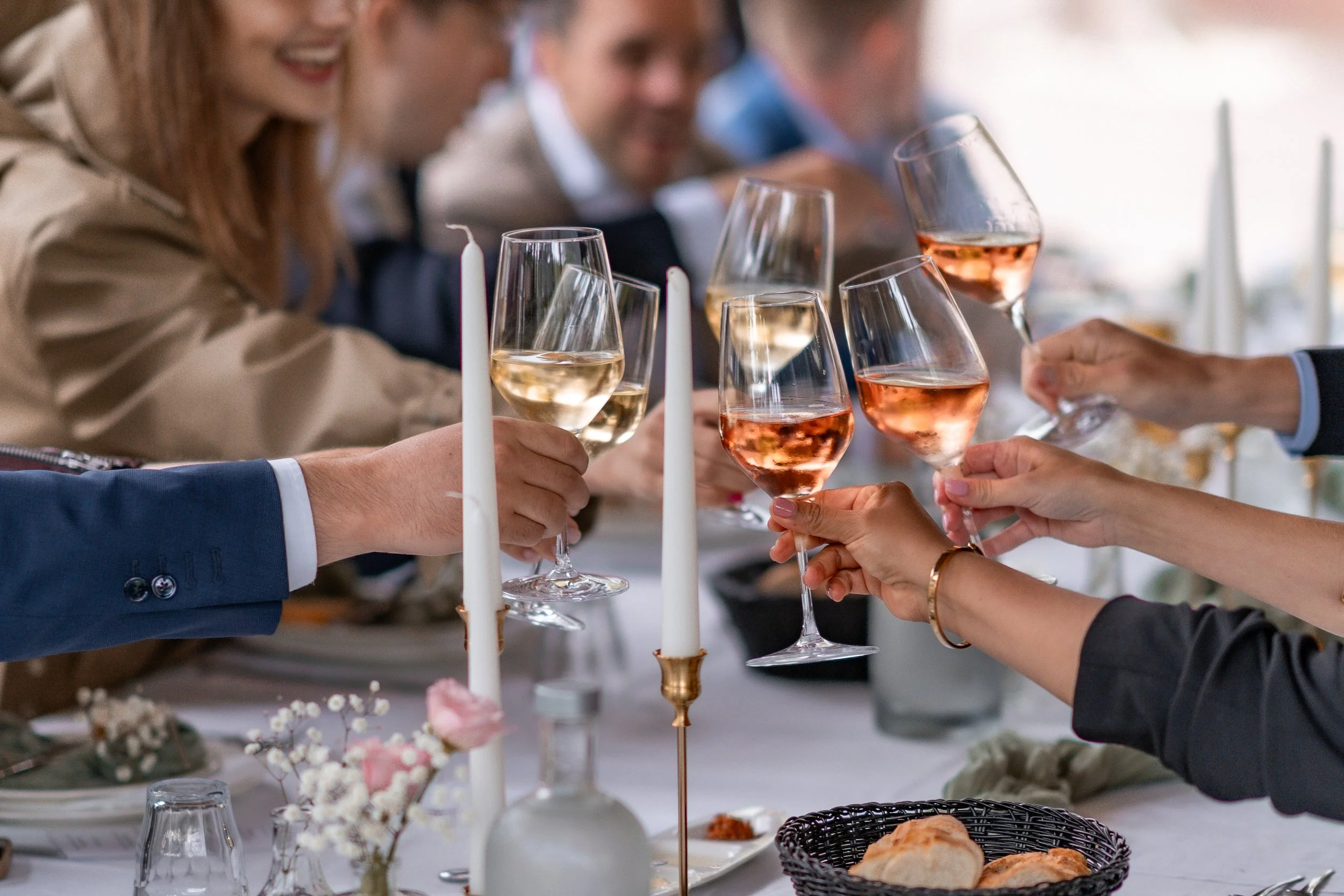 People at a celebration raising glasses of white and rosé wine in a toast, with candles, flowers, bread, and tableware on the table.