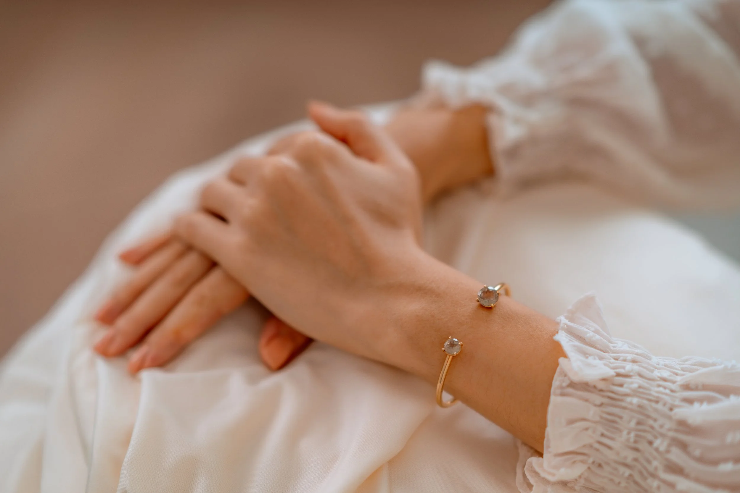 Close-up of a woman's hand resting on her lap, adorned with a gold bracelet with two round gemstones and a white ruffled sleeve.