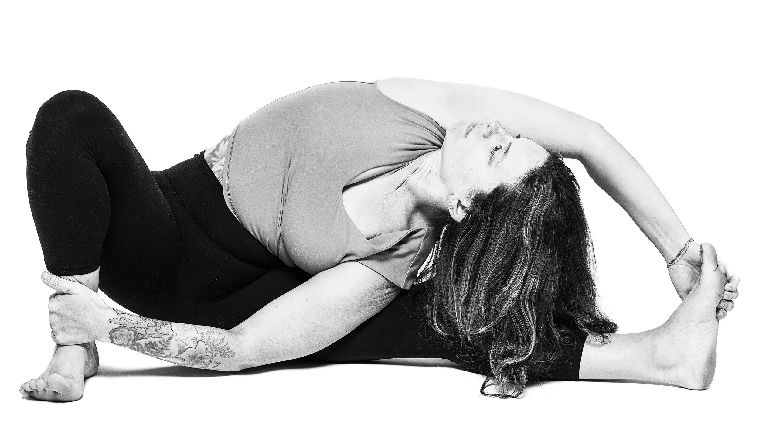A woman performing a yoga pose with her left arm wrapped around her back and her right hand holding her left foot, lying on a white surface.