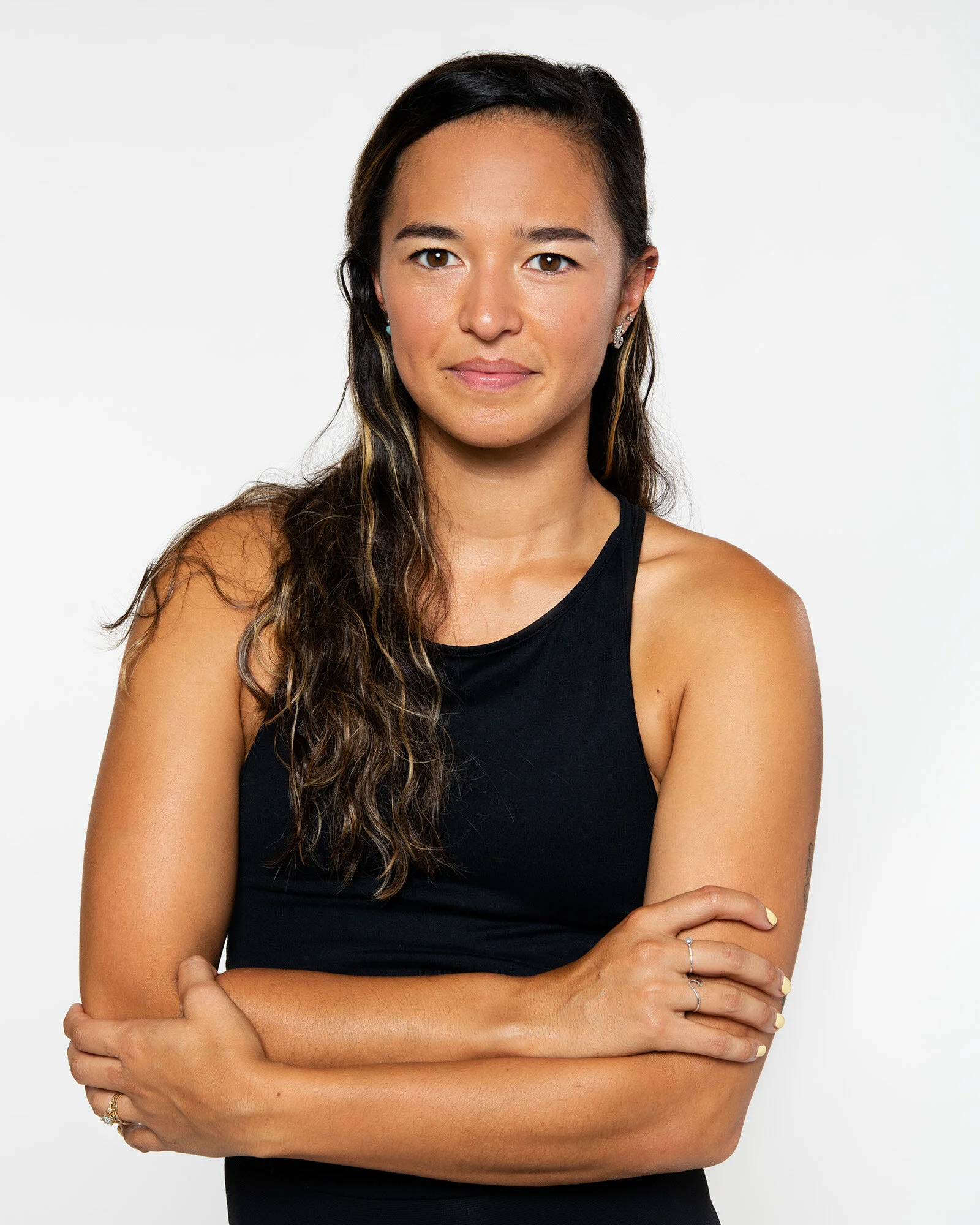 A woman with long, wavy brown hair wearing a black sleeveless top standing with her arms crossed against a plain white background.