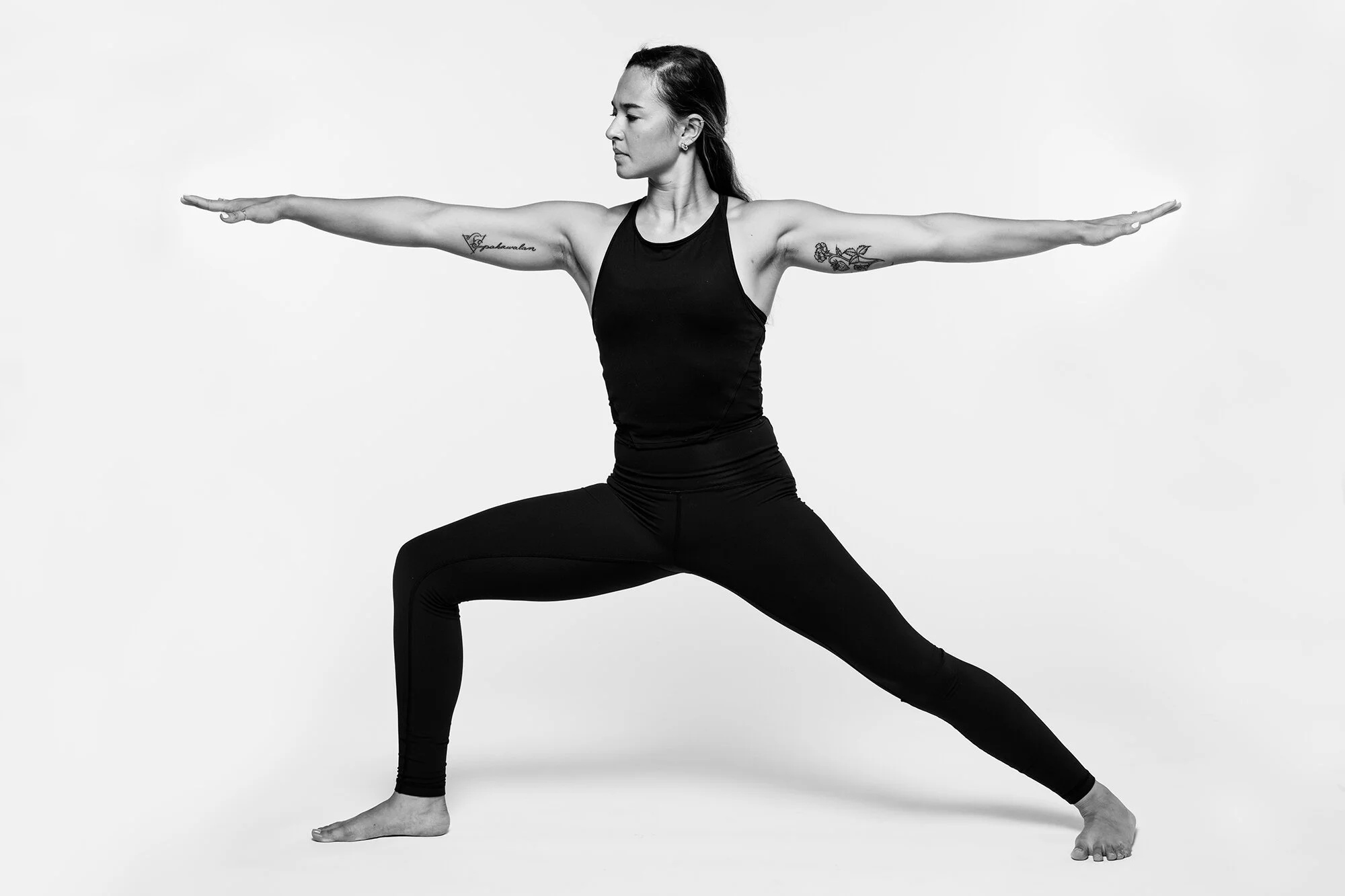 A woman in a black athletic outfit practicing yoga in a Warrior II pose against a plain background.