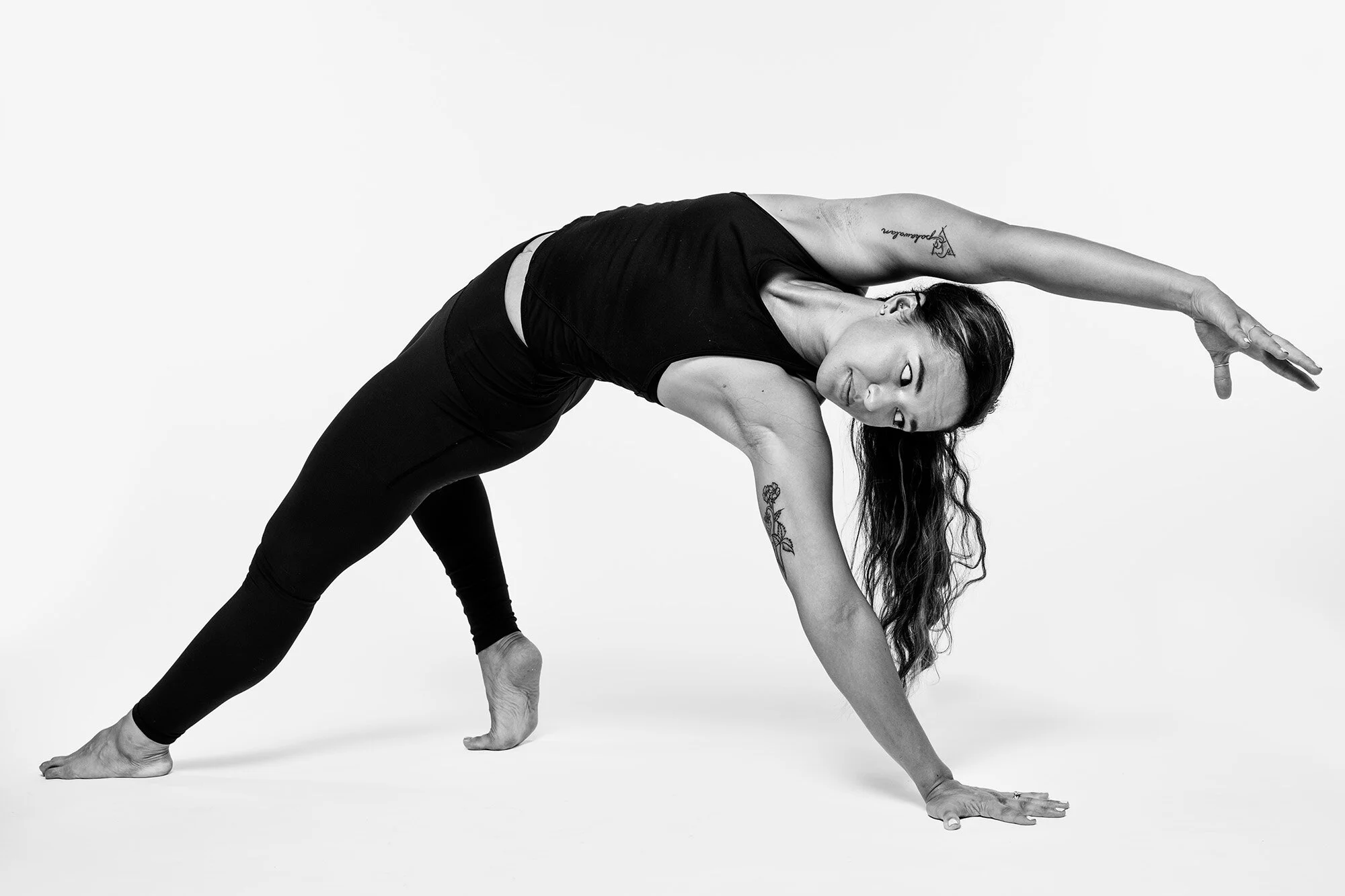 A woman doing a yoga pose in black clothing against a plain white background, with tattoos on her arms and long hair hanging down.