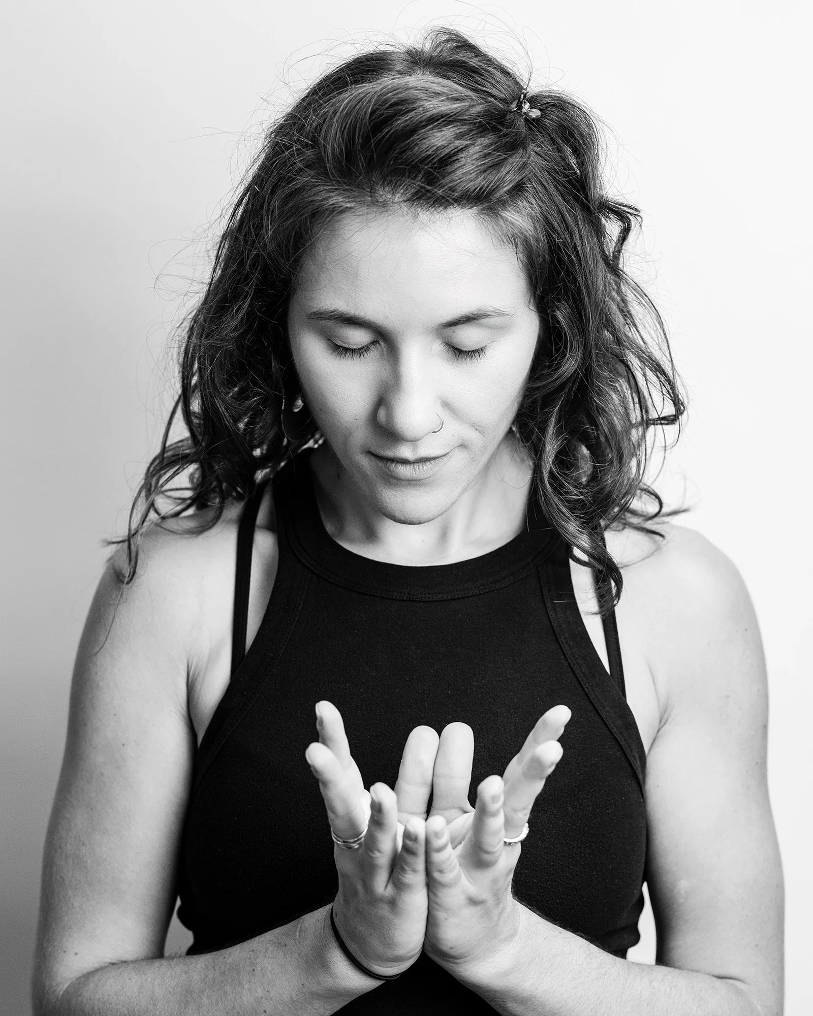 A woman in a black sleeveless top with curly hair and nose piercing stands with her eyes closed and hands clasped together in a prayer position.