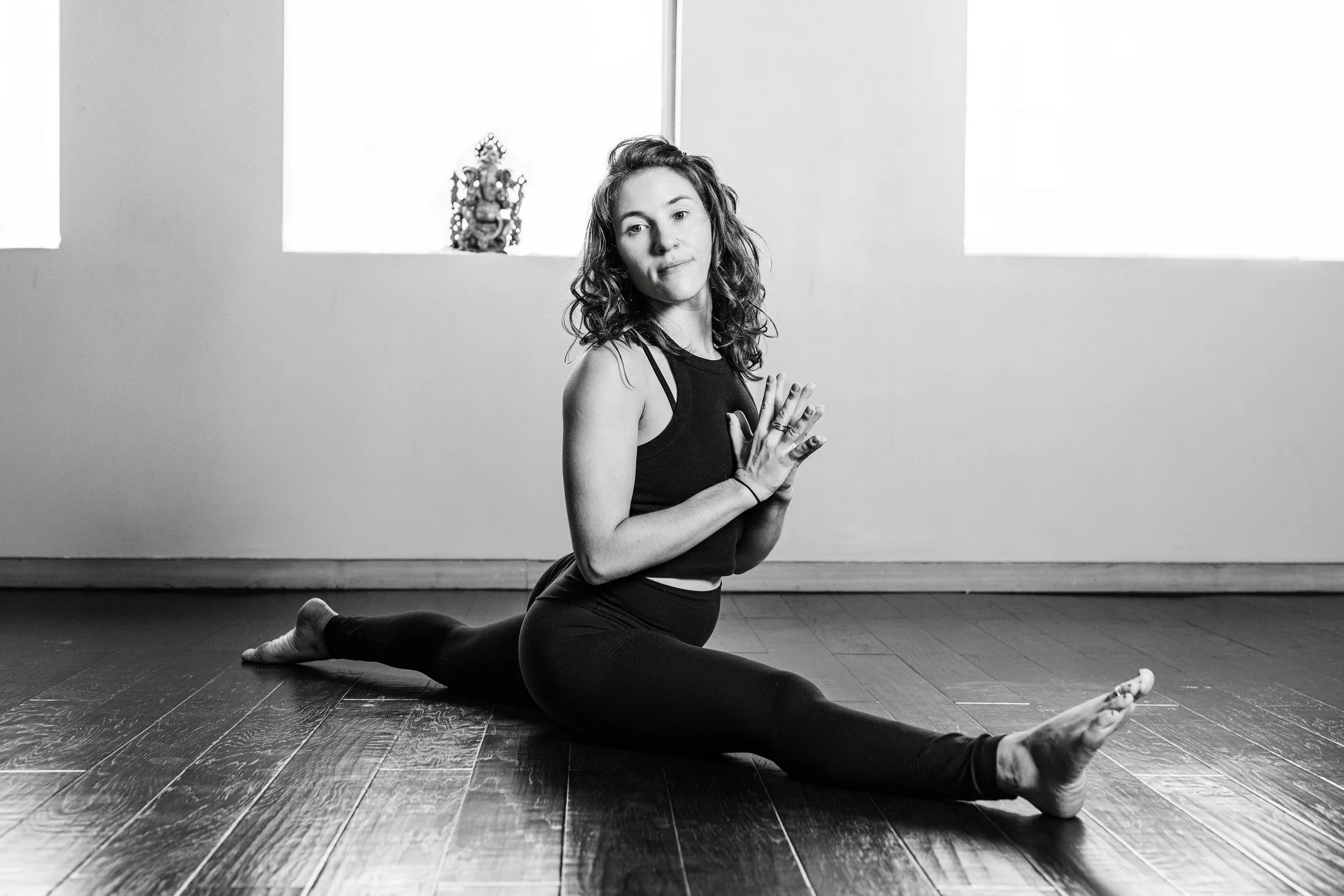 A woman practicing yoga indoors, sitting on the floor in a split position with hands pressed together in a prayer position. She has curly hair and is wearing a sleeveless top and leggings. There are two windows in the background with natural light coming through.