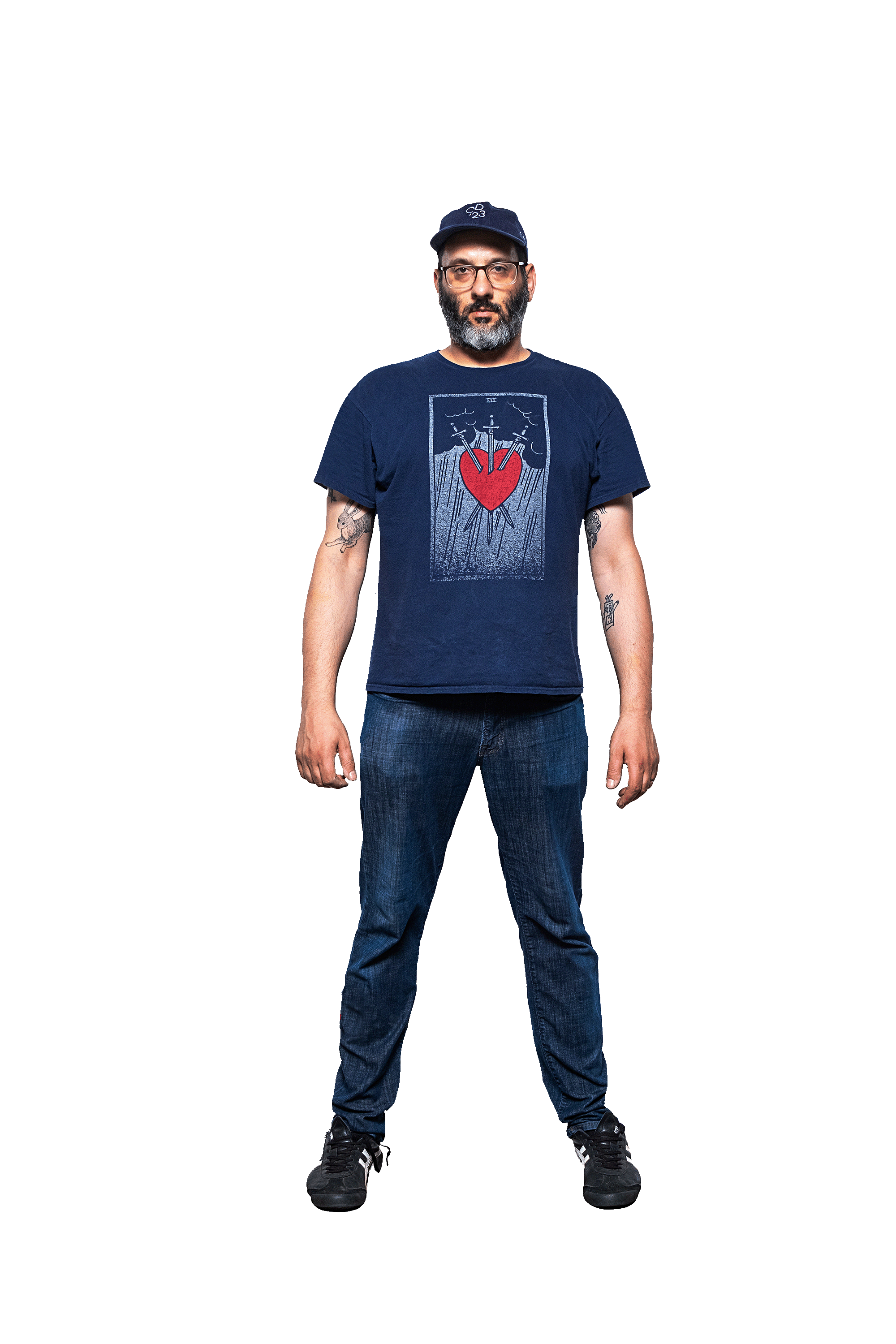 A man standing against a black background, wearing a navy blue t-shirt with a graphic of a red heart and swords, jeans, sneakers, a cap, and glasses.