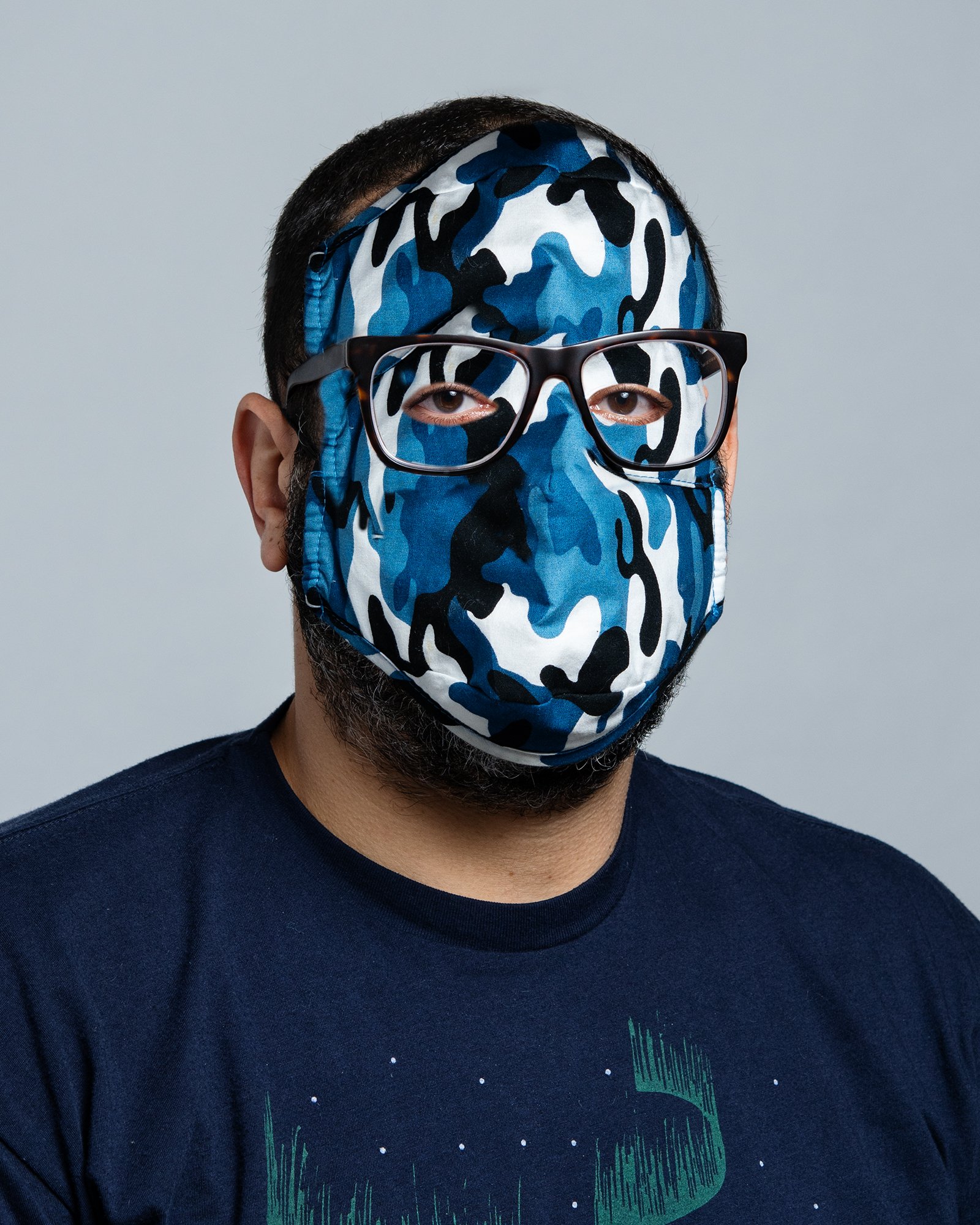 A man wearing a camouflage face mask, glasses, and a dark t-shirt against a plain background.