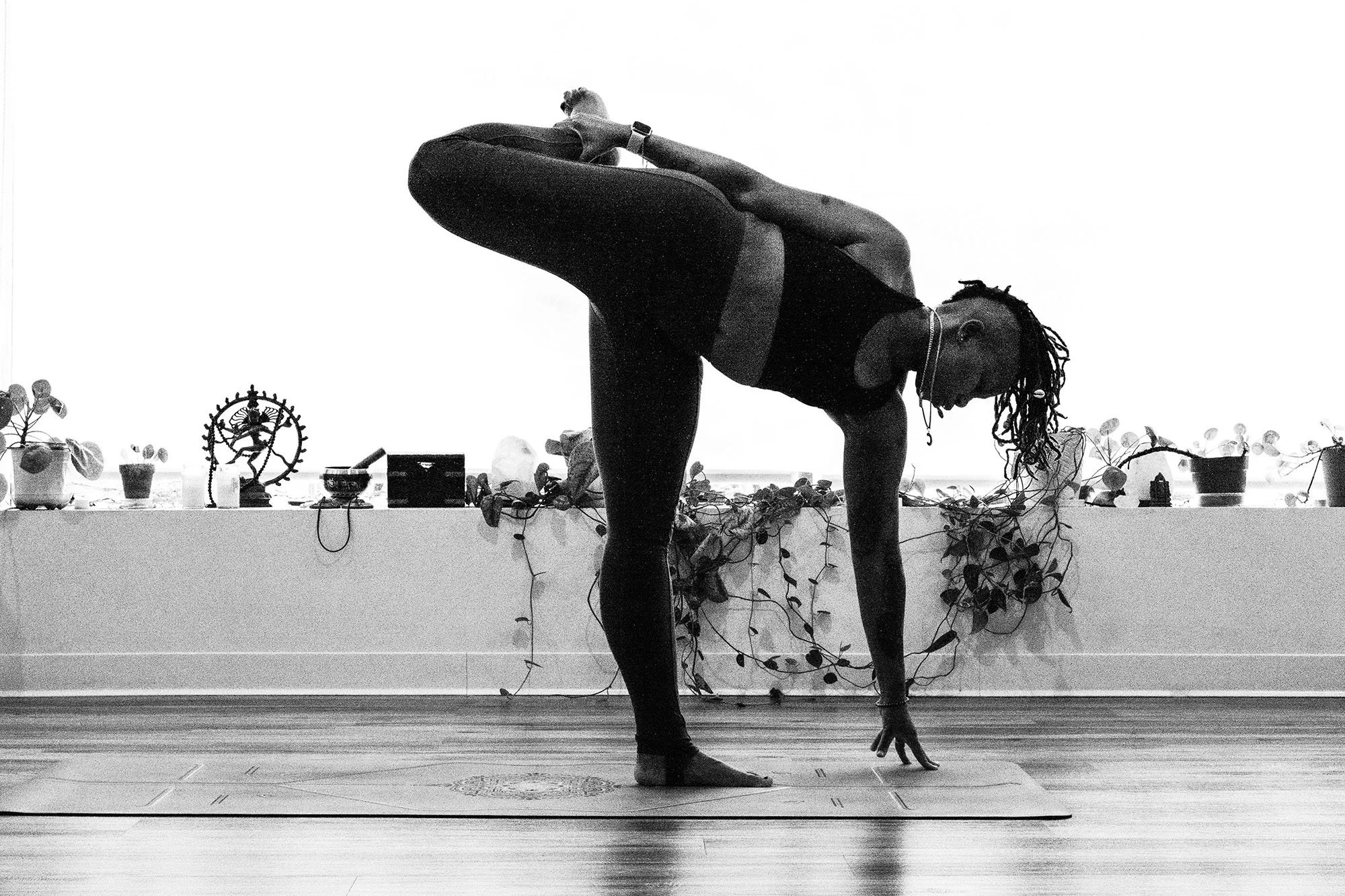 A woman practicing yoga indoors, balancing on one leg with her other leg bent behind her and her hand holding her foot, while extending her other arm downward, on a yoga mat.