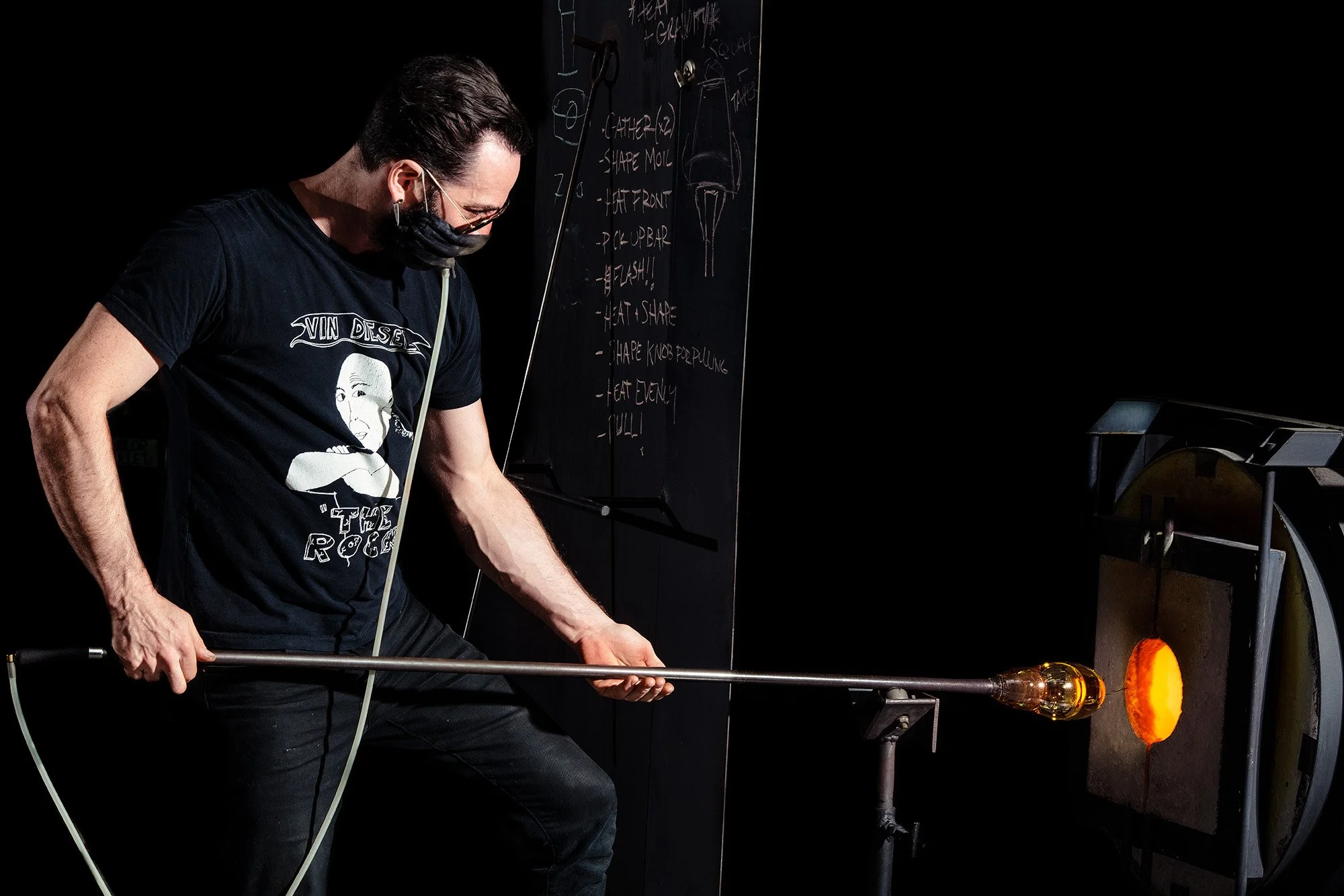 A glassblower wearing a black t-shirt and mask working with molten glass at a furnace in a black-walled studio.