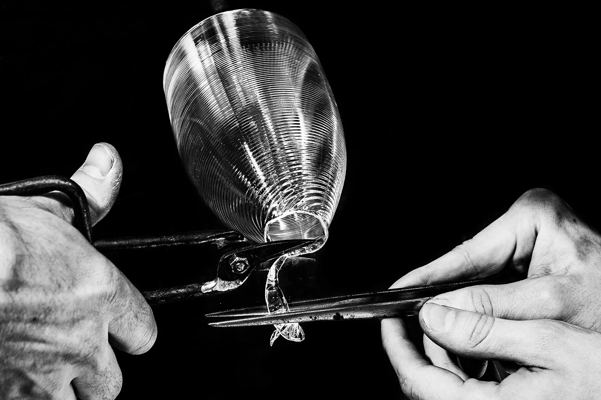 Close-up of a person’s hands using pliers to handle a metallic shell, with another hand holding the shell on a black background.