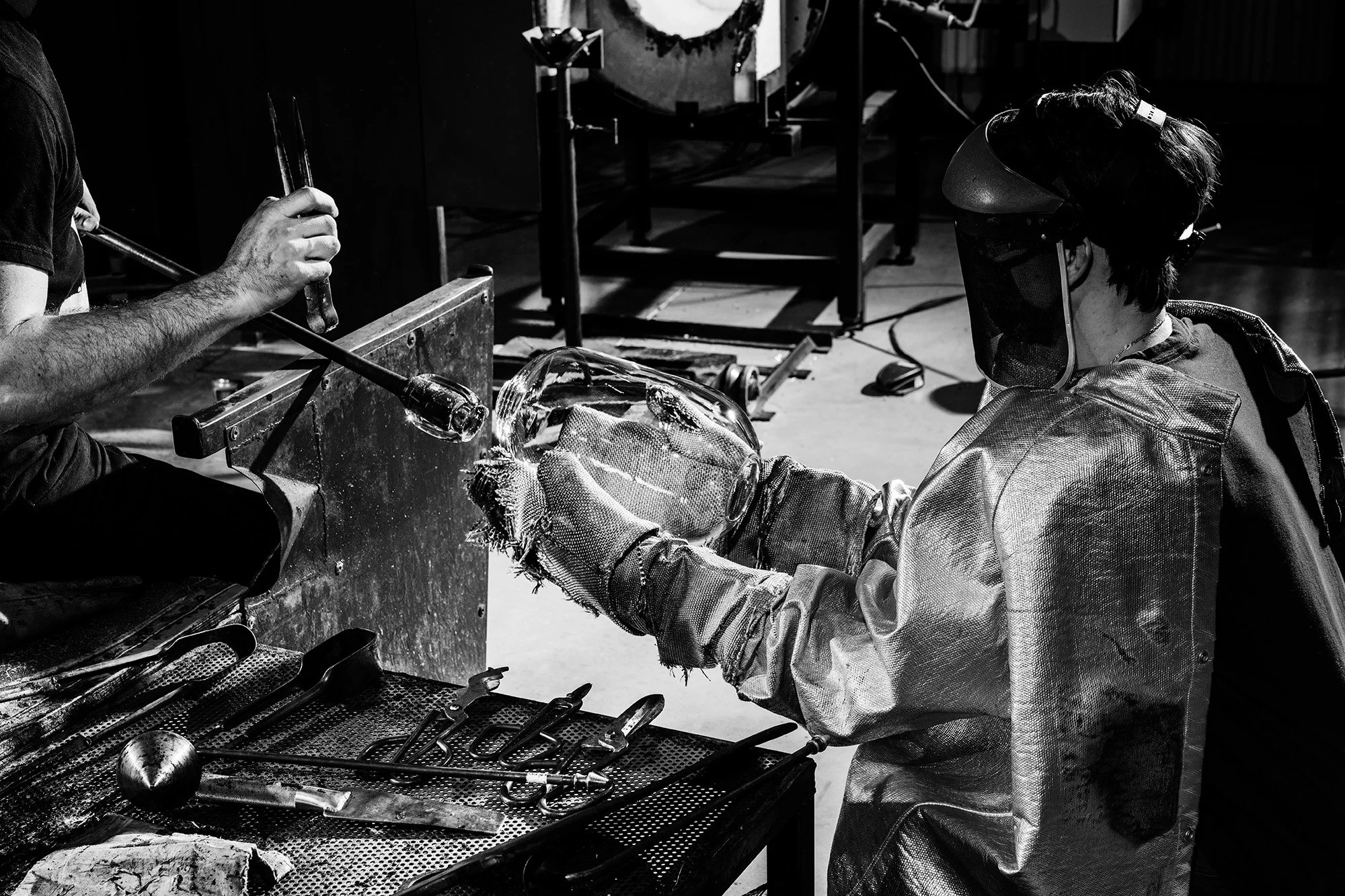 A glassblower wearing protective gear including a face shield and gloves working on shaping glass in a workshop, with tools and equipment on the workbench.