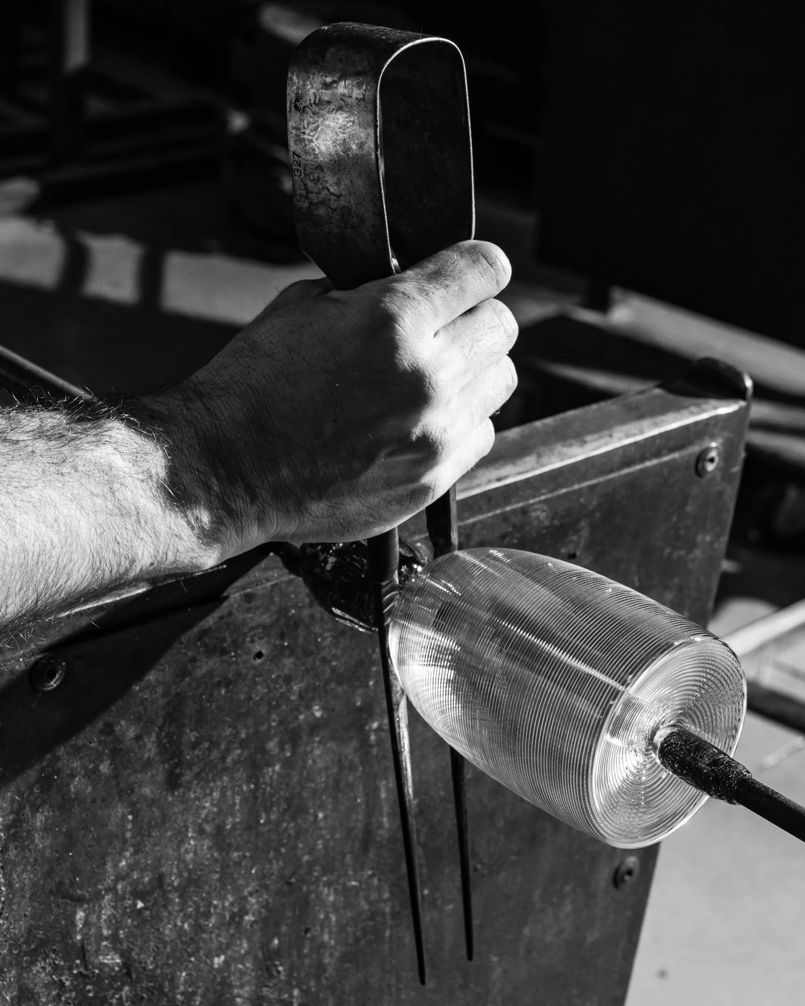 A black and white photo of a person's hand holding a metal tool, working on a transparent glass object in a workshop setting.