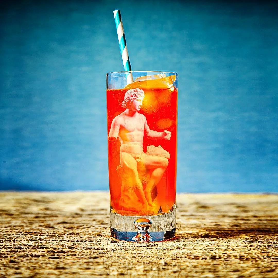 A tall glass of red-colored beverage with ice cubes, a lemon slice, and a striped straw, featuring a mini sculpture of a seated, muscular male figure from classical sculpture inside the drink, with a wooden surface and blue background.