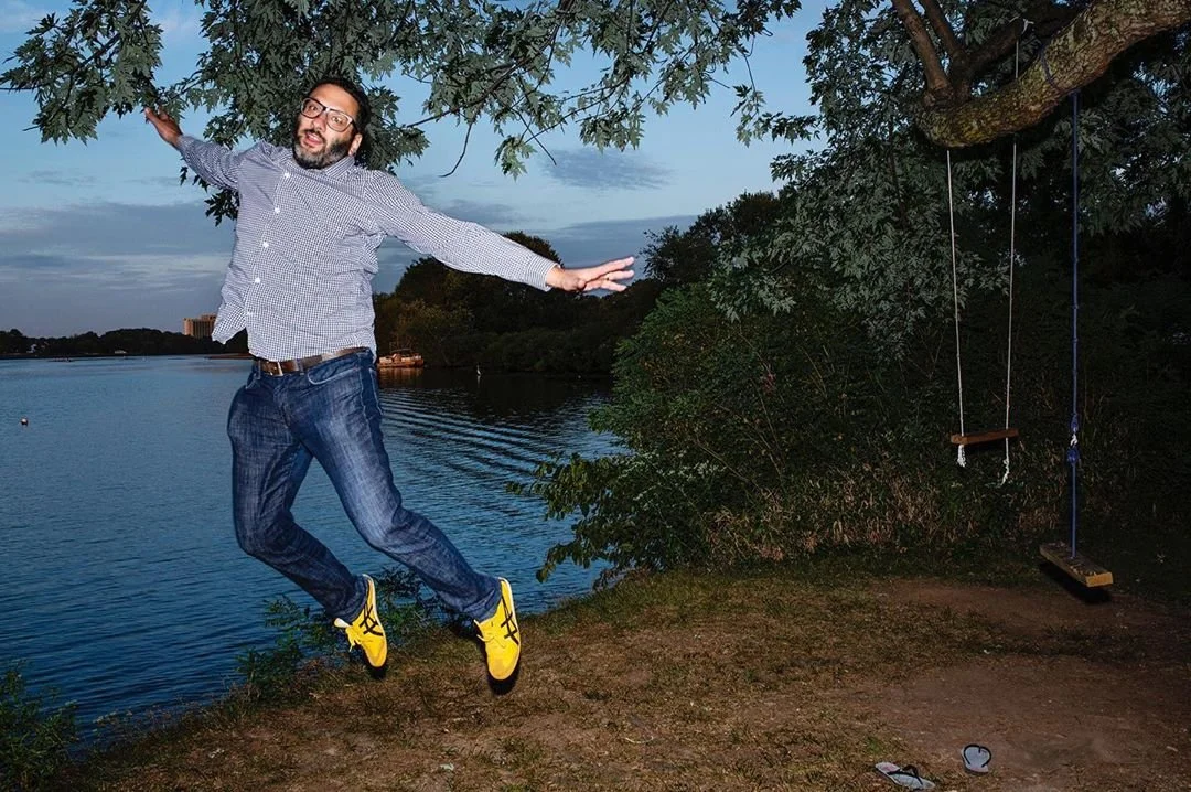A man wearing glasses, a checkered shirt, and jeans is jumping near a lakeside with a tree and a swing in the background during evening or dusk.