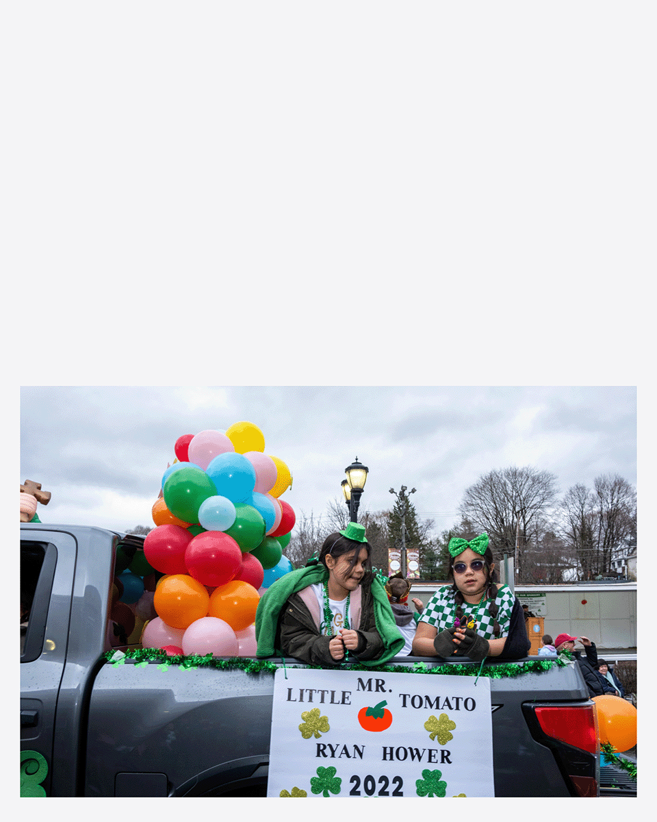 Two girls riding in a parade float decorated with green, orange, and pink balloons, wearing green accessories and sunglasses, with a sign that reads "Mr. Little Tomato Ryan Hower 2022" and shamrock decorations.