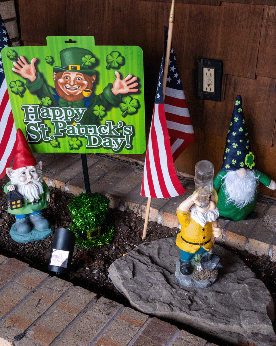 Decorative St. Patrick's Day display with a sign reading 'Happy St. Patrick's Day!' featuring leprechaun graphics, small American flags, and garden gnome figurines dressed as leprechauns, set against a brick wall.