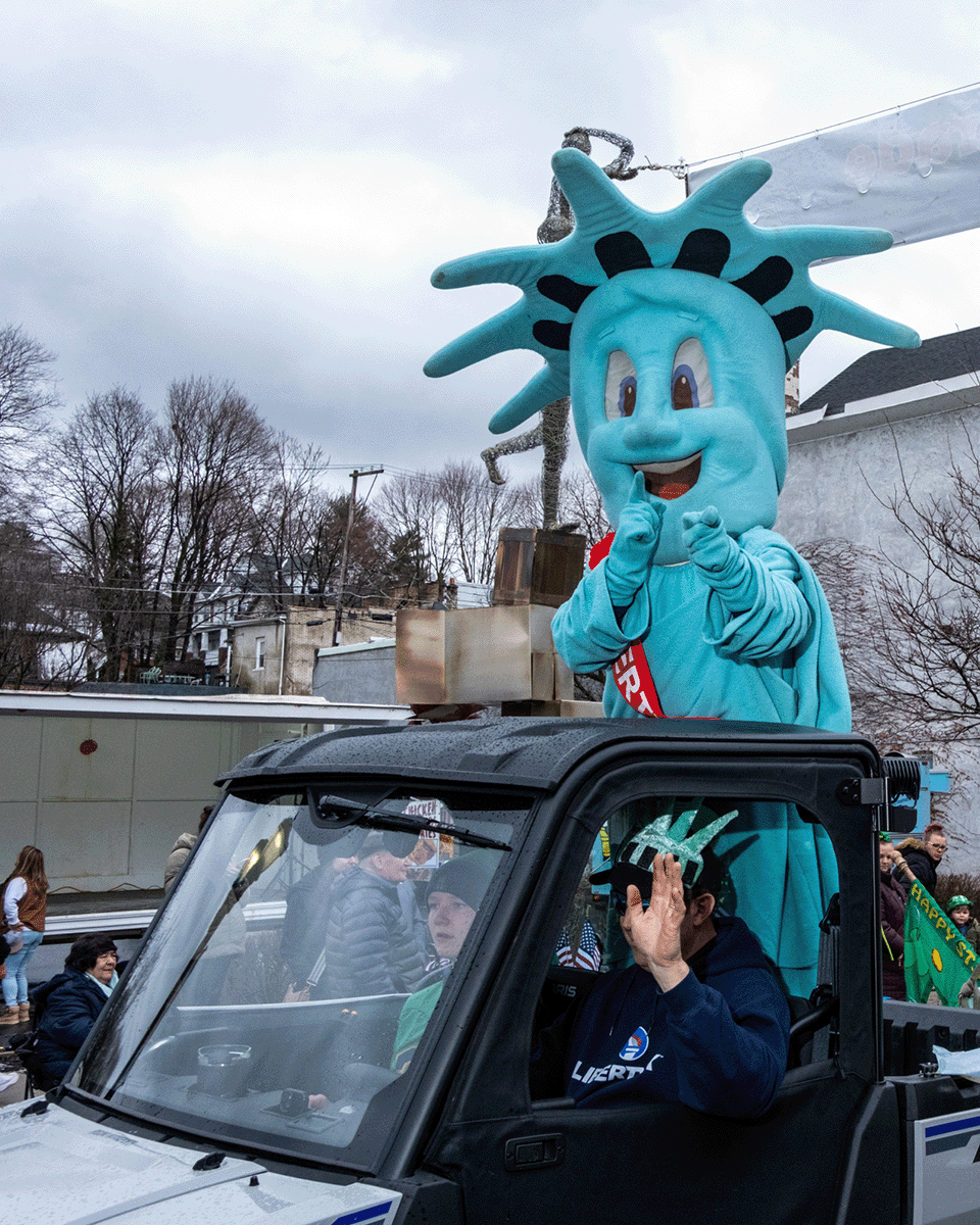 A person dressed in a Statue of Liberty costume standing on a parade float, waving and smiling. The float is being driven by a person wearing a Liberty University hoodie.