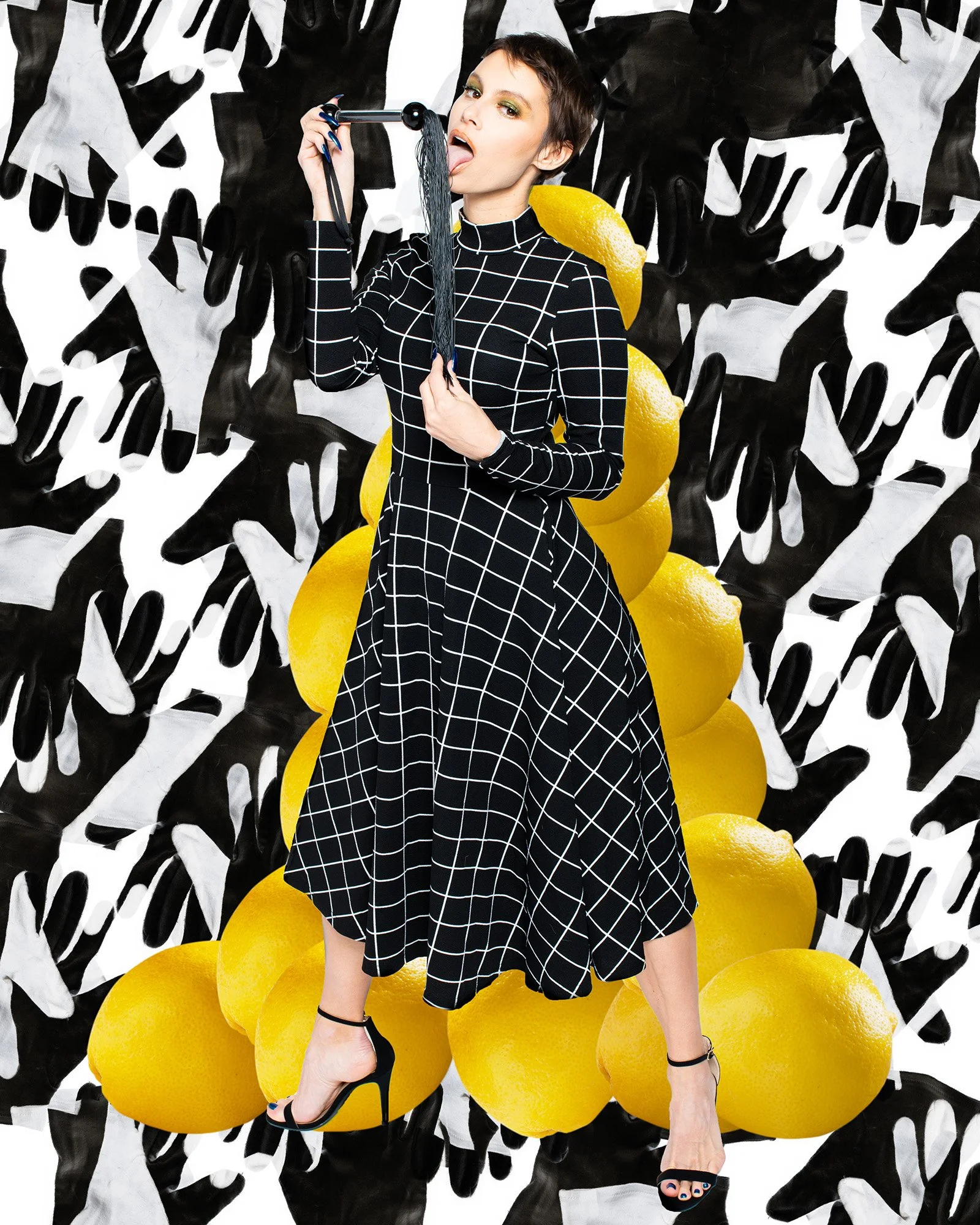 A woman in a black dress with white grid patterns poses against a surreal background of lemons and monochrome handprints, holding a black hairbrush to her tongue.